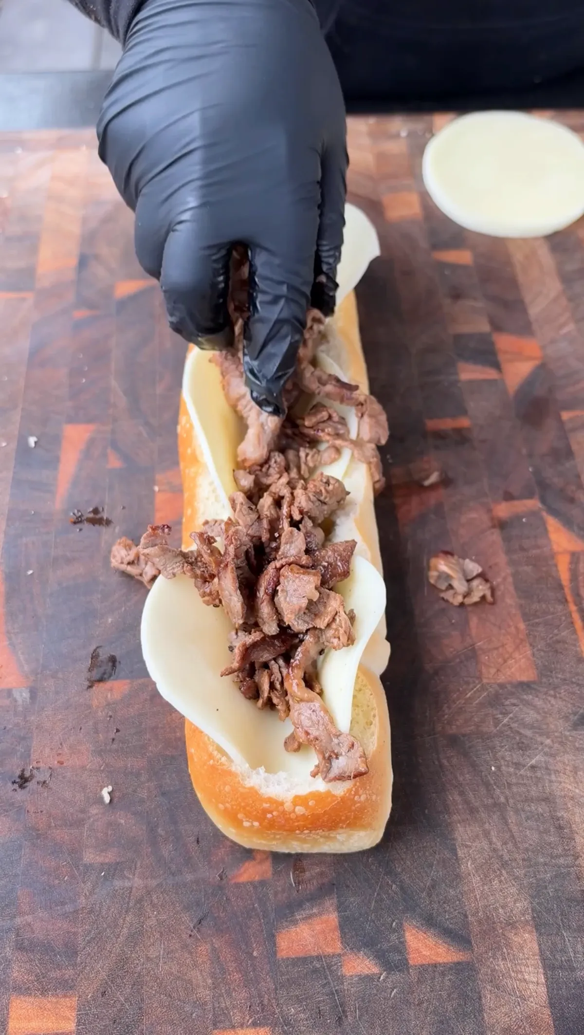 Gloved hand placing thinly sliced ribeye strips onto provolone-lined French baguette on a wooden cutting board