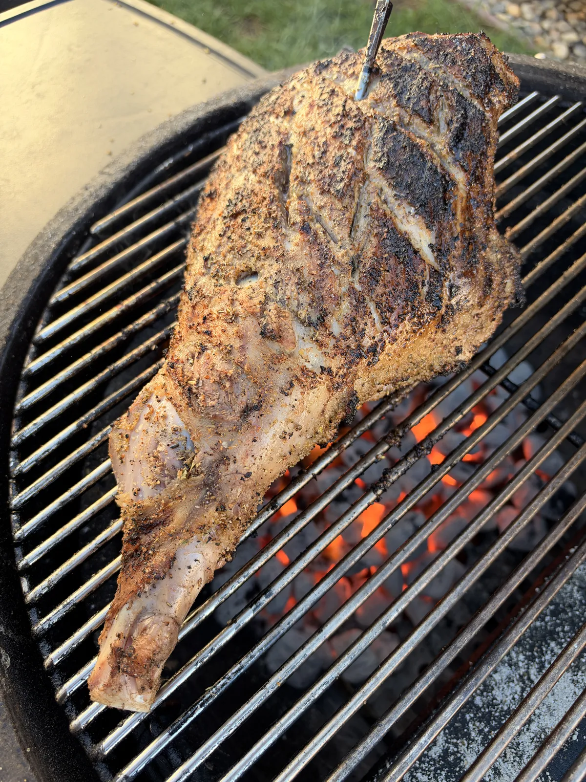 Seasoned leg of lamb searing on hot grill grates with glowing charcoal, developing a deep-brown crust