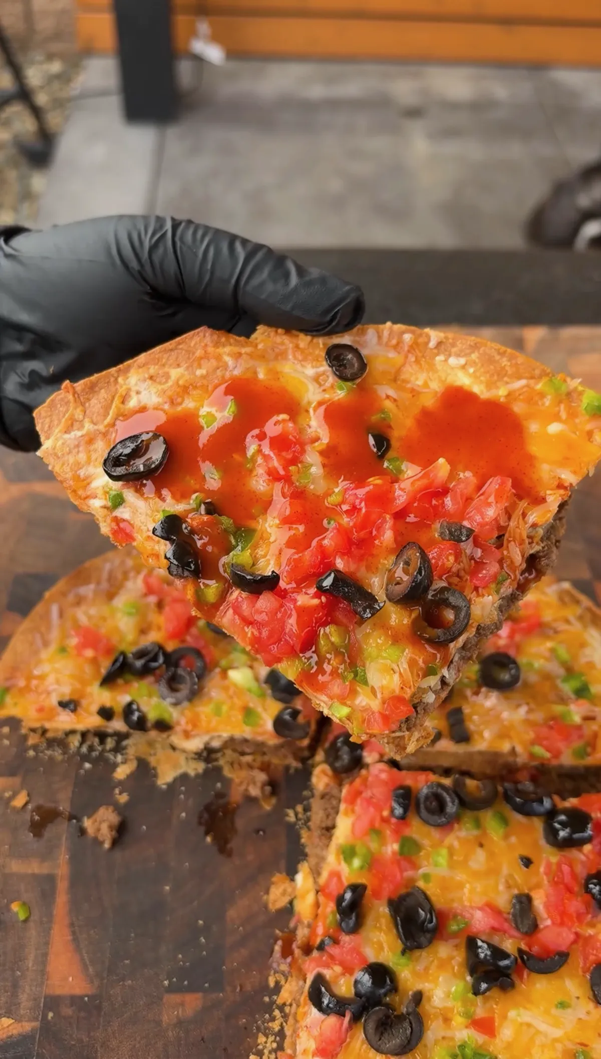 Gloved hand lifting a slice of Mexican pizza with Taco Bell taco sauce, tomatoes, olives and melted Colby Jack