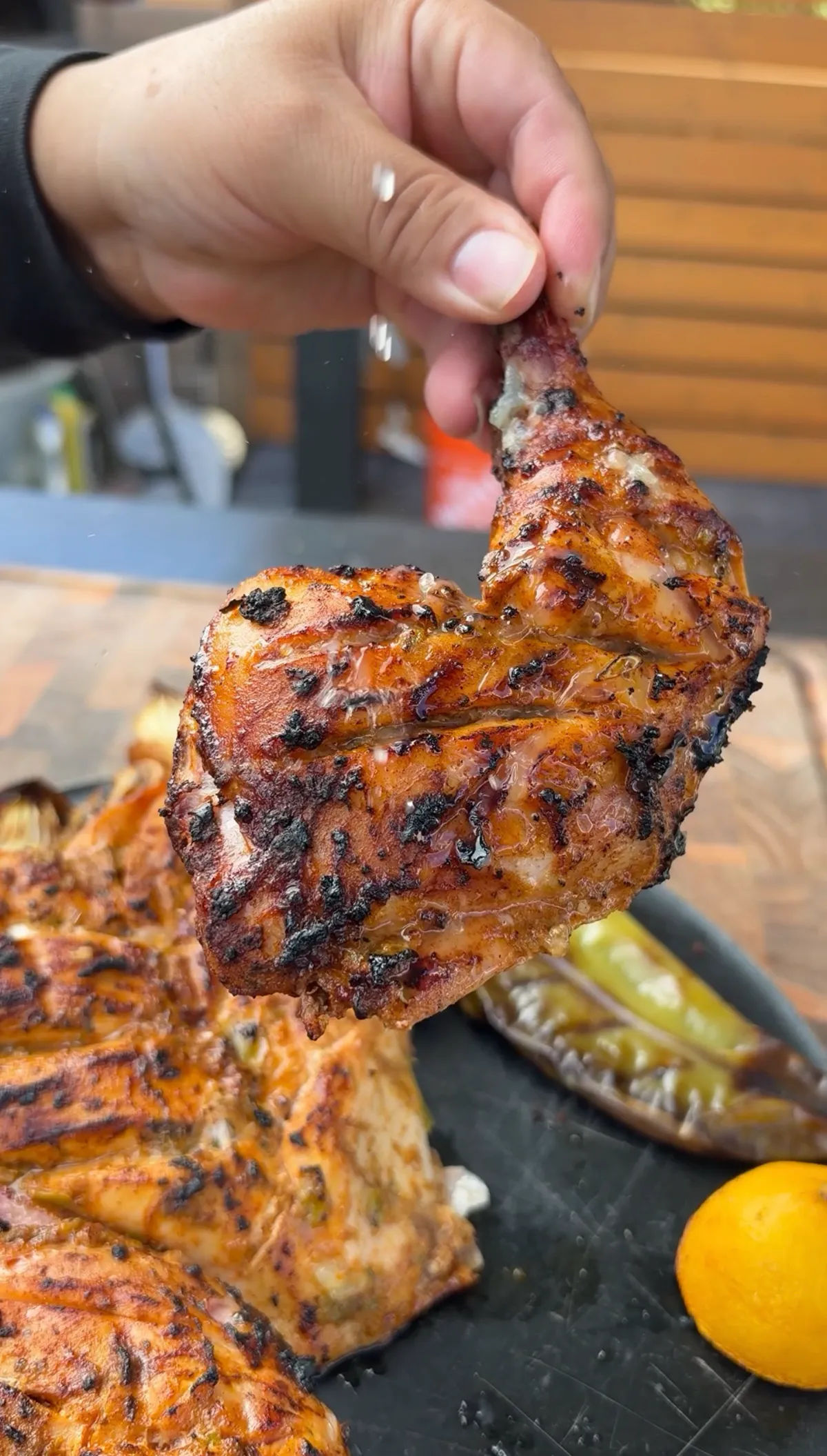 Hand lifting a juicy grilled chicken quarter with charred chipotle smoked paprika crust over a platter with roasted Anaheim pepper and lemon