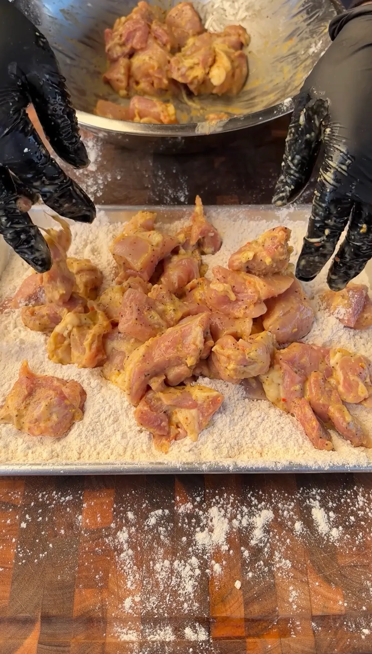 Hands in black gloves transferring lemon pepper marinated chicken thigh pieces onto a flour-dusted baking tray for coating