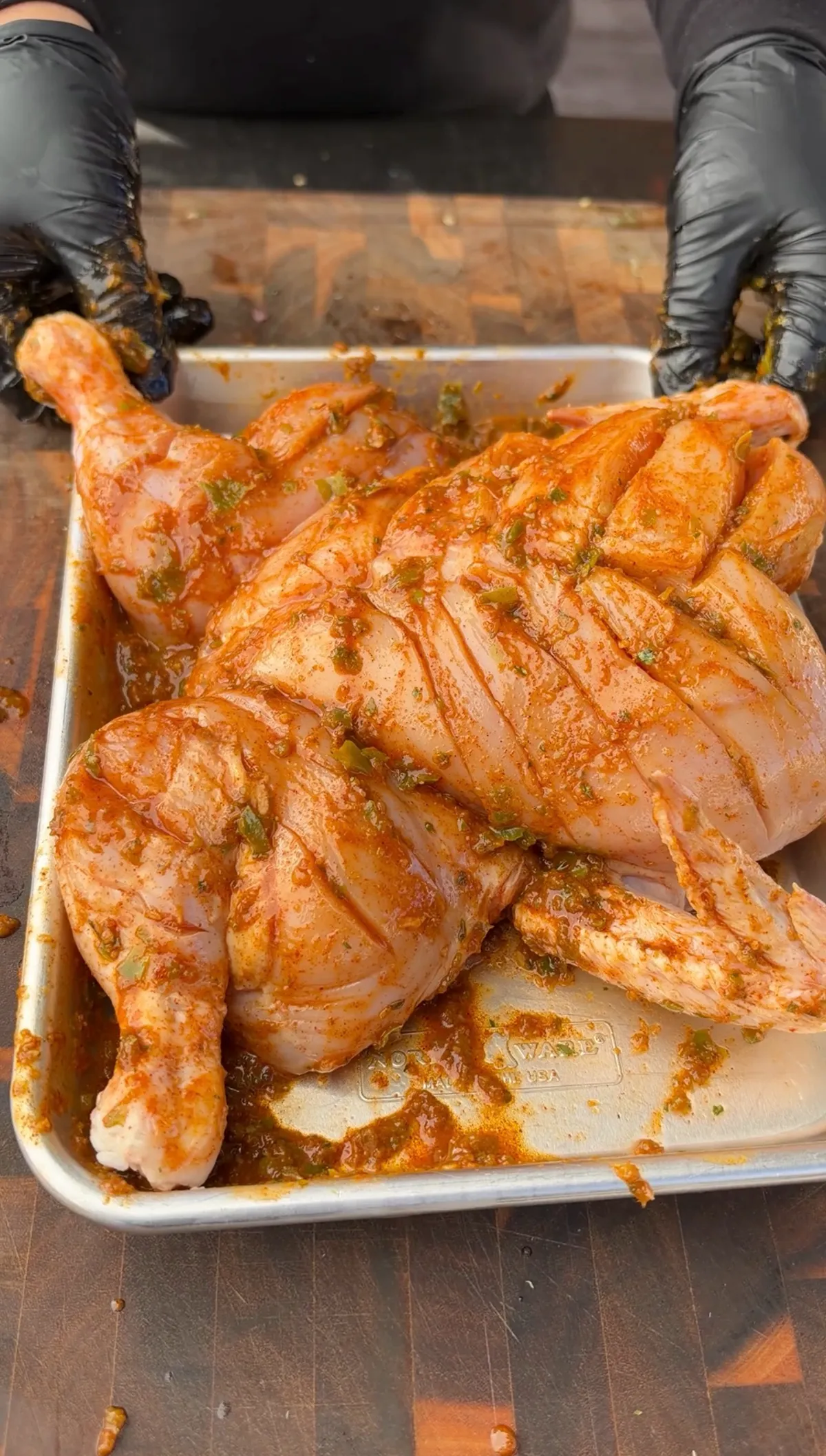 Hands in black gloves holding a sheet pan with scored, spatchcocked chicken coated in green chile lime marinade