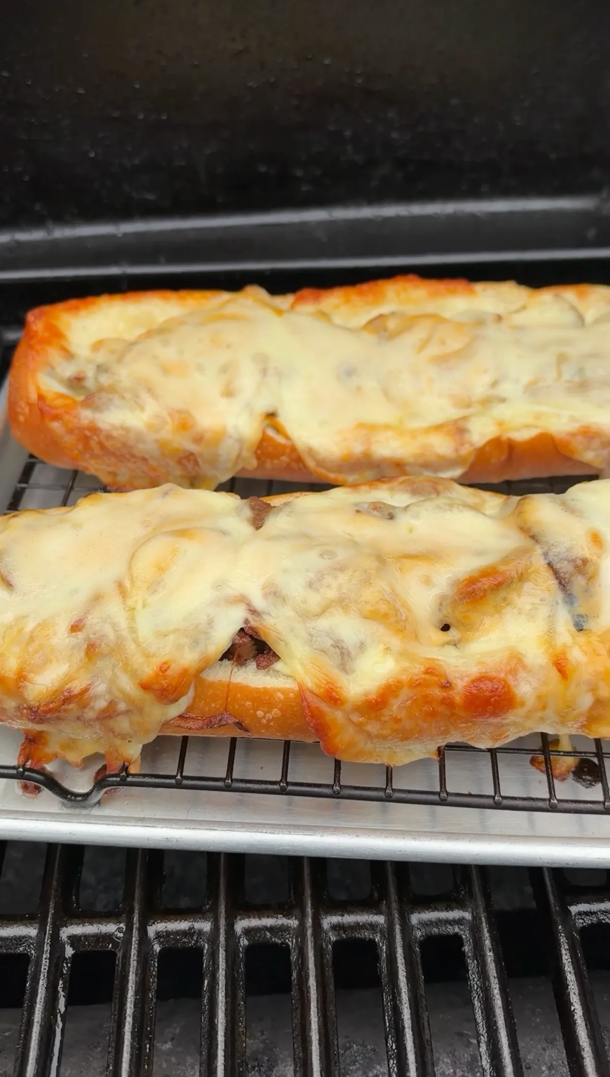 Two cheesesteak stuffed baguette halves with bubbling melted provolone baking on a wire rack inside a grill at 350°F