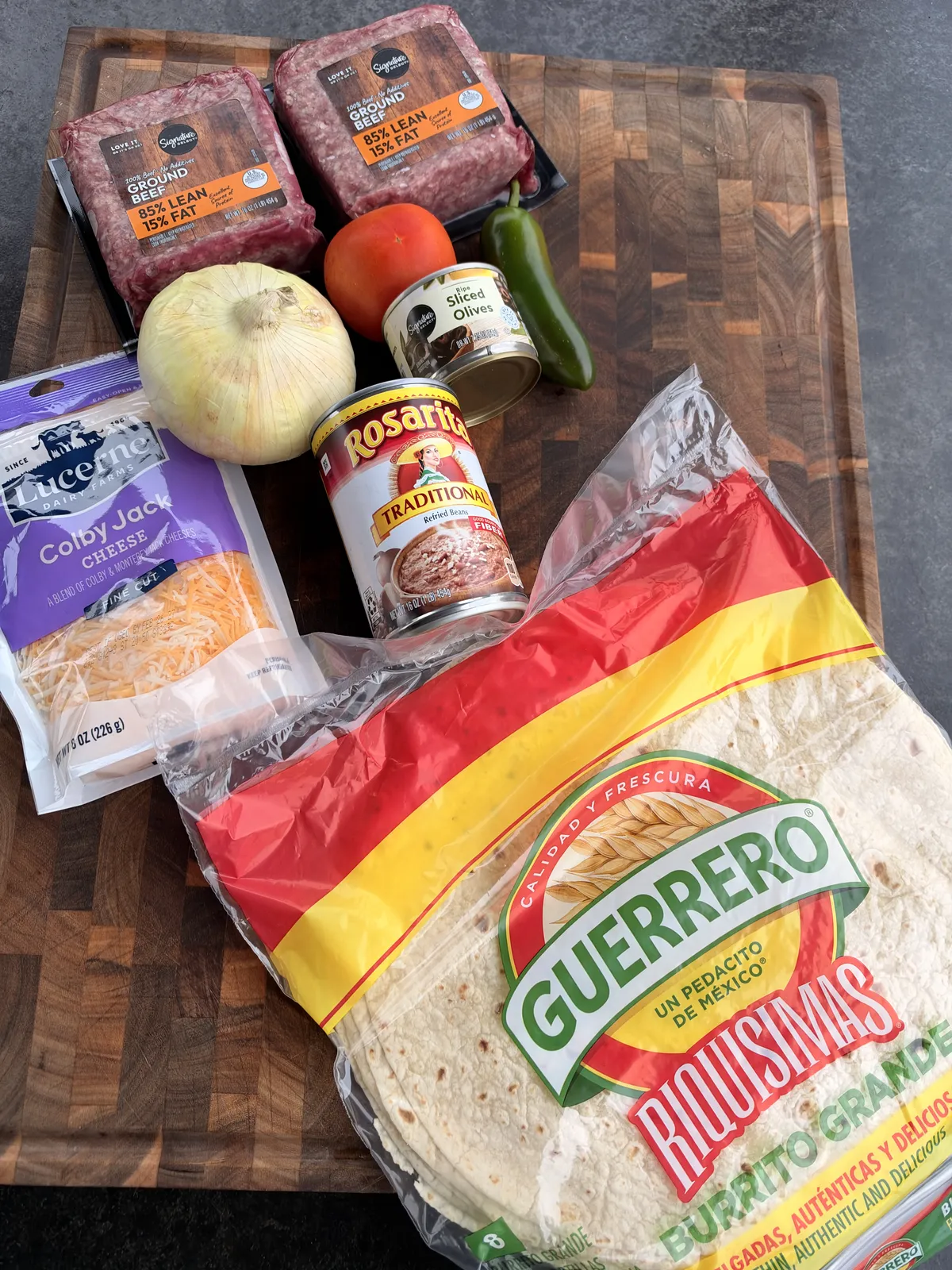 Taco Bell copycat Mexican pizza ingredients: ground beef, refried beans, flour tortillas, Colby Jack cheese, tomato, jalapeño, and sliced black olives on a wooden cutting board