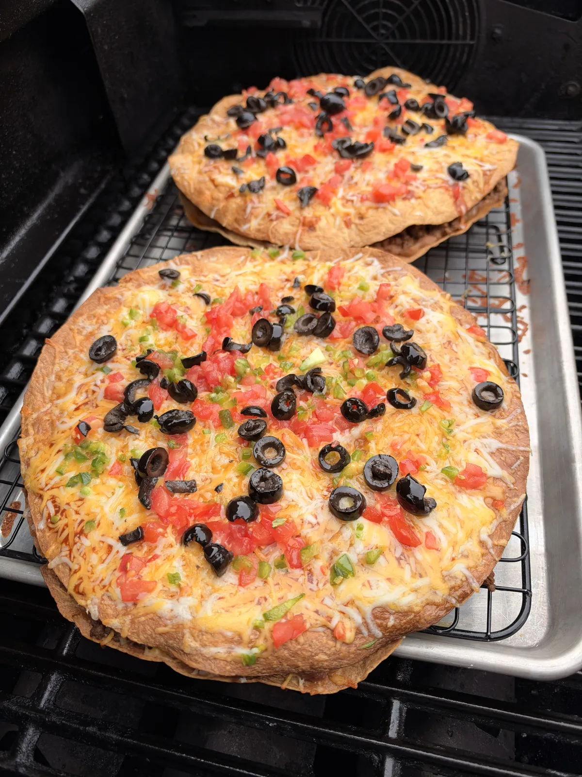 Two homemade Taco Bell copycat Mexican pizzas with melted Colby Jack cheese, diced tomatoes, sliced black olives, and jalapeños on a wire rack inside a baking sheet on a grill.