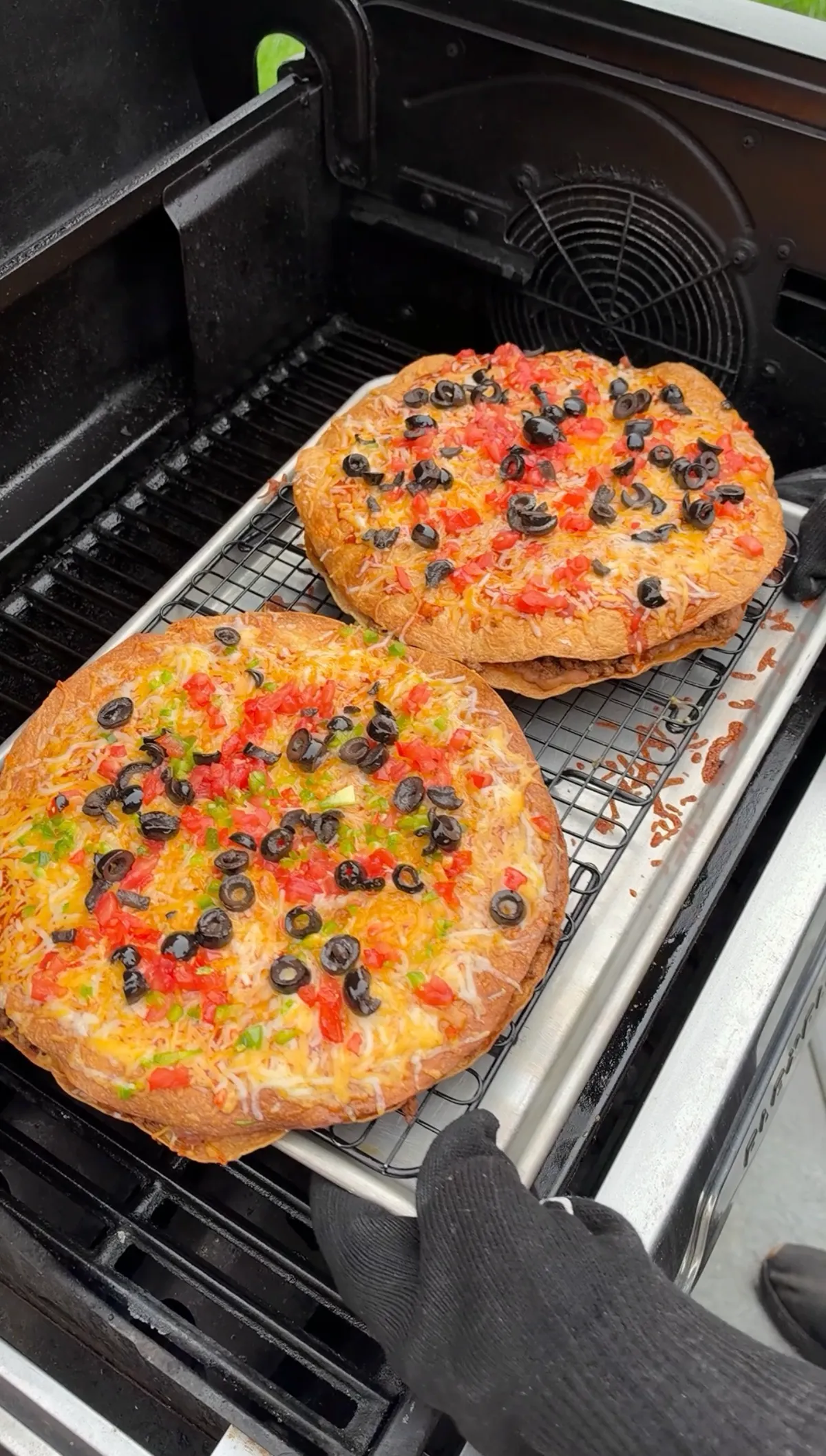 Two assembled Mexican pizzas with melted Colby Jack cheese, black olives, diced tomatoes and jalapeños on a wire rack inside the grill