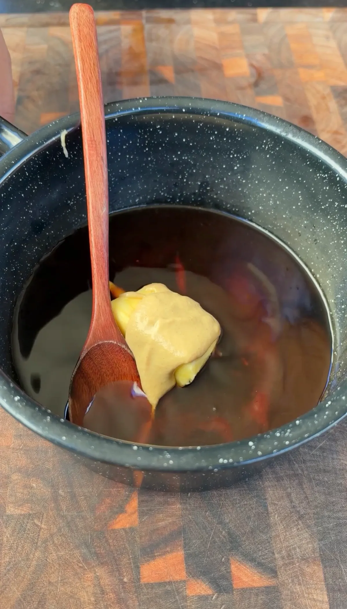 A dollop of yellow mustard on a wooden spoon resting in a dark BBQ sauce in a speckled black saucepan