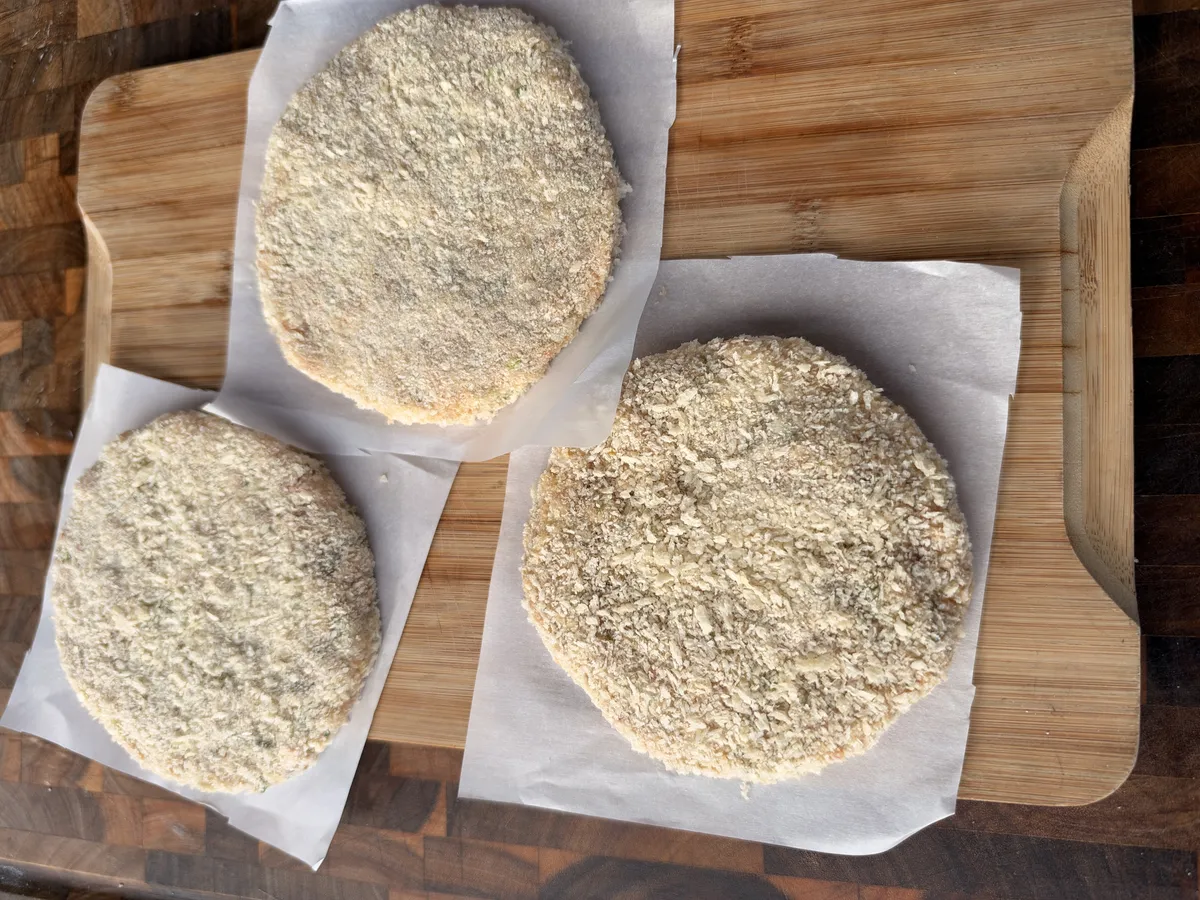 Three raw shrimp burger patties coated in panko breadcrumbs on parchment paper, ready to fry