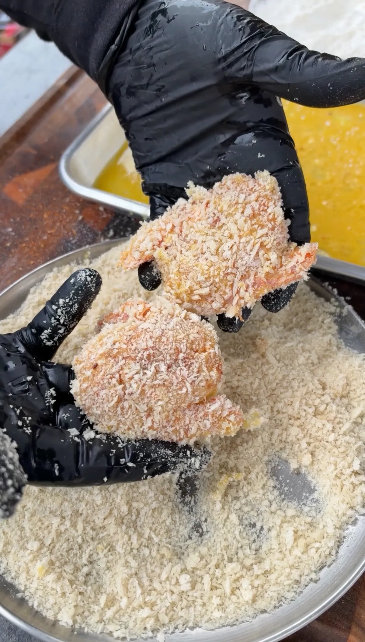 Gloved hands pressing large raw shrimp into panko breadcrumbs over a metal bowl, with egg wash visible nearby