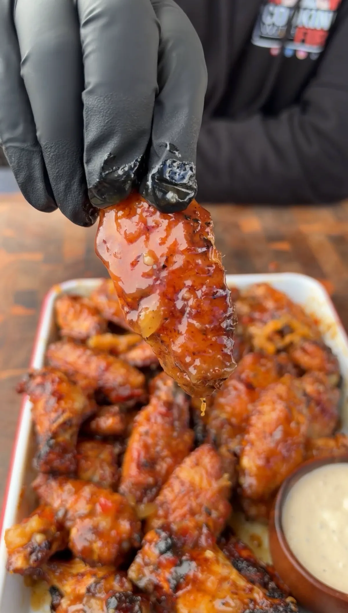Gloved hand holding a sticky pineapple habanero glazed chicken wing over a tray of wings with ranch dipping sauce