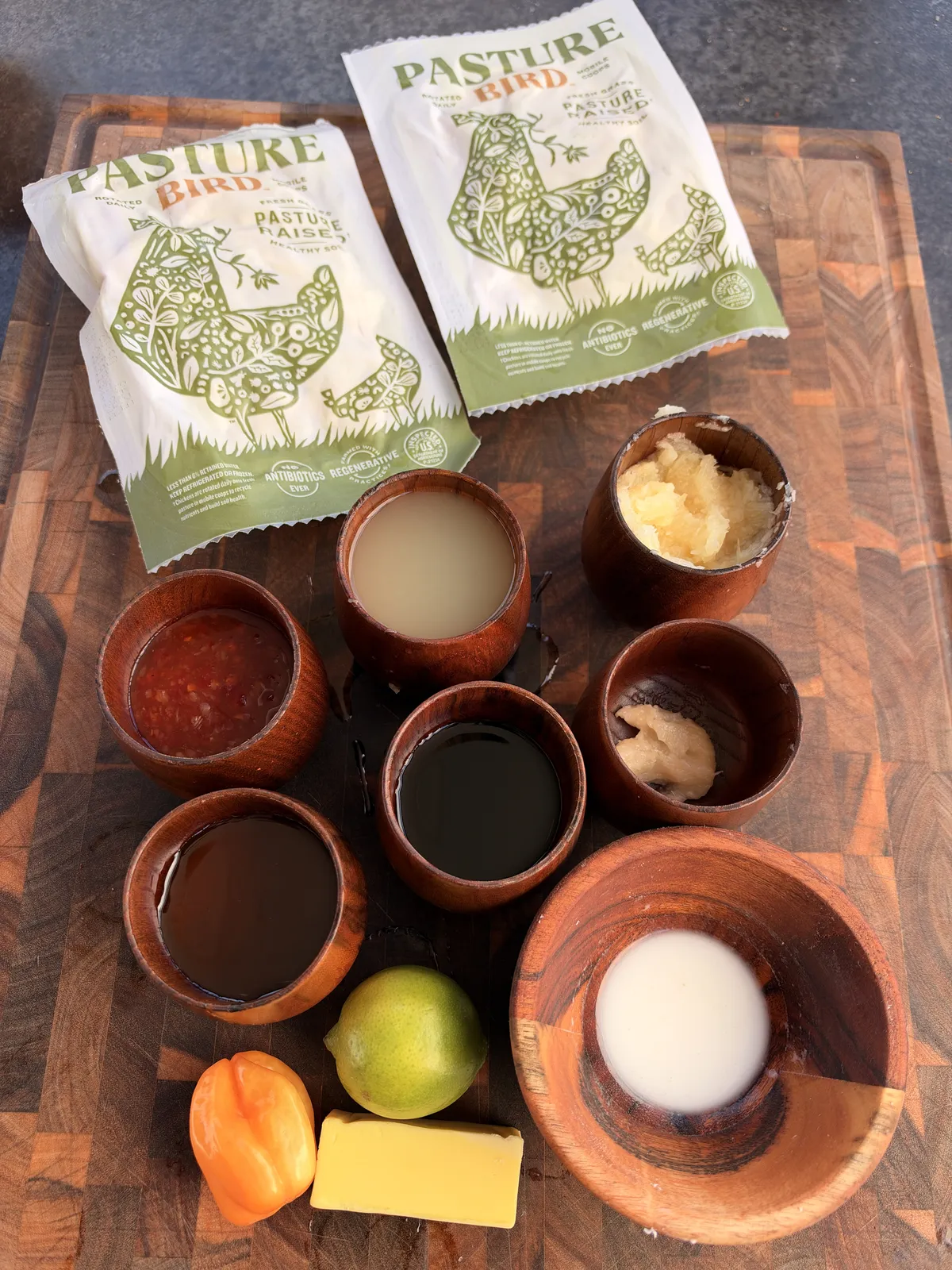 Pineapple habanero grilled chicken wing ingredients: Pasture Bird wings, sweet chili sauce, soy sauce, honey, pineapple juice, crushed pineapple, minced garlic, habanero pepper, lime, butter, and cornstarch slurry arranged in wooden bowls on a cutting board