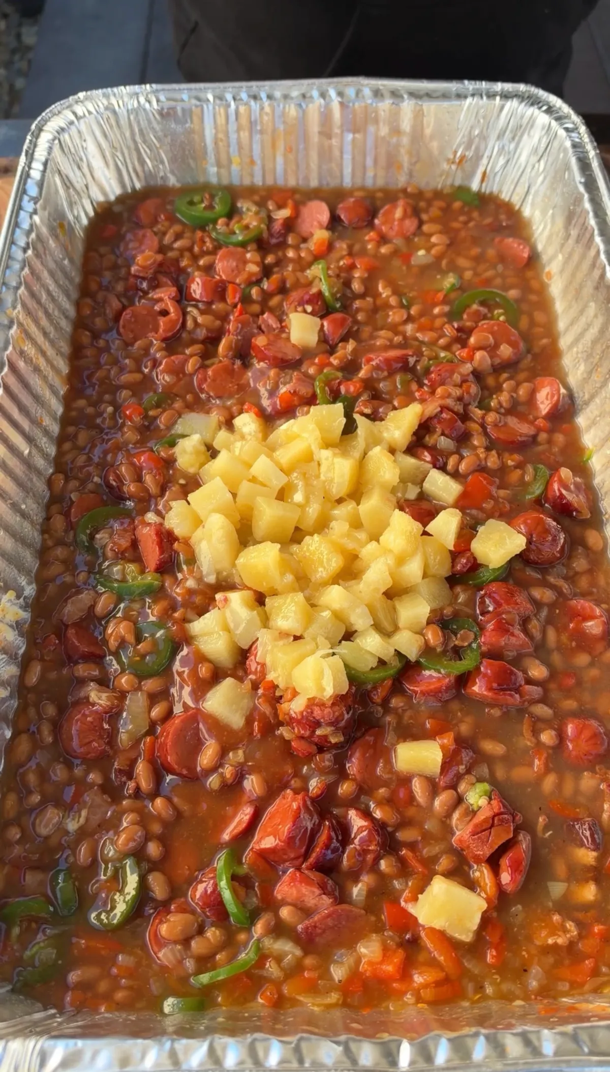 Smoked BBQ baked beans in a foil pan topped with drained pineapple tidbits, jalapeño rounds, and sliced hot dogs