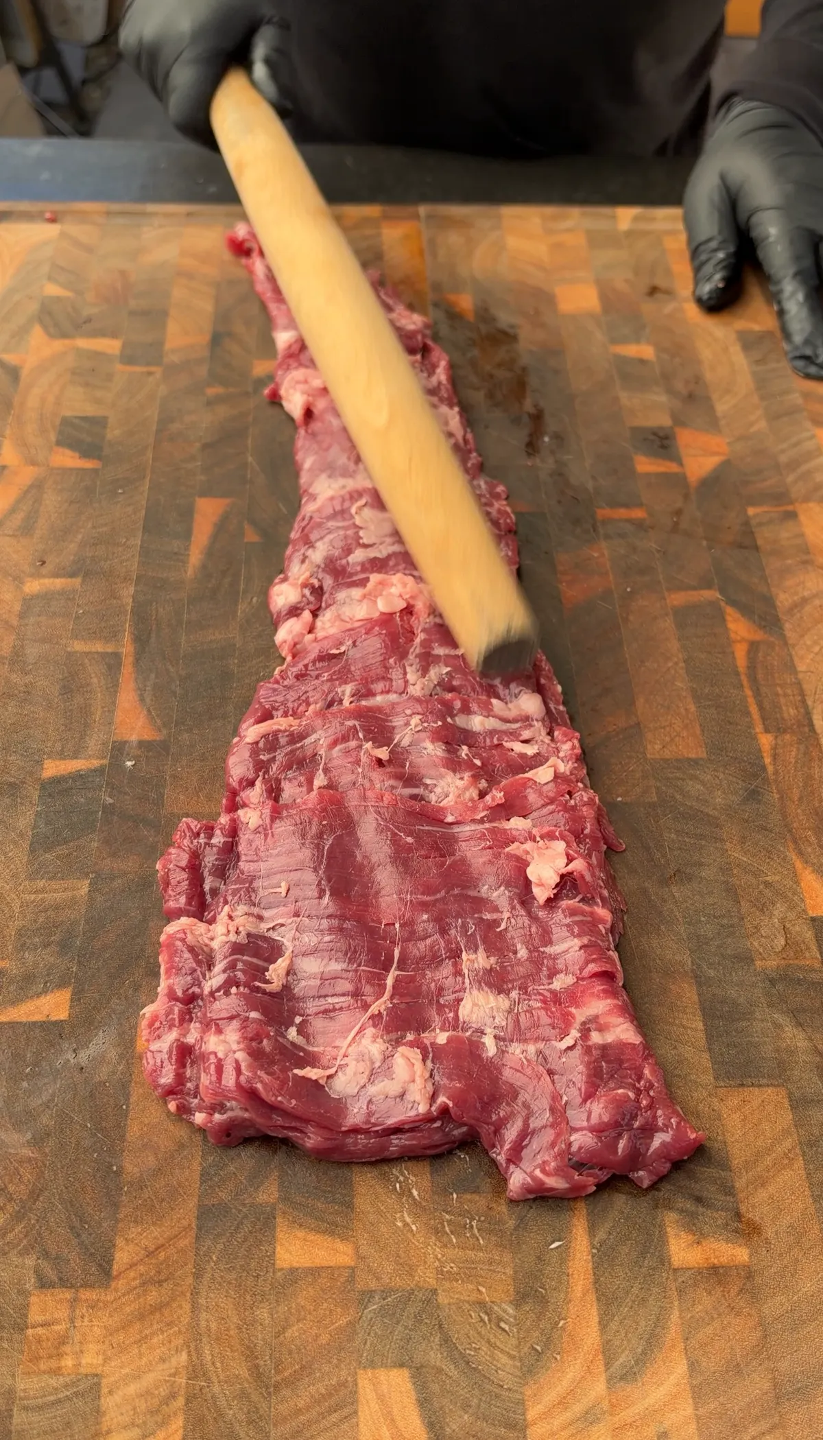 Skirt steak being flattened with a wooden rolling pin on a butcher block cutting board
