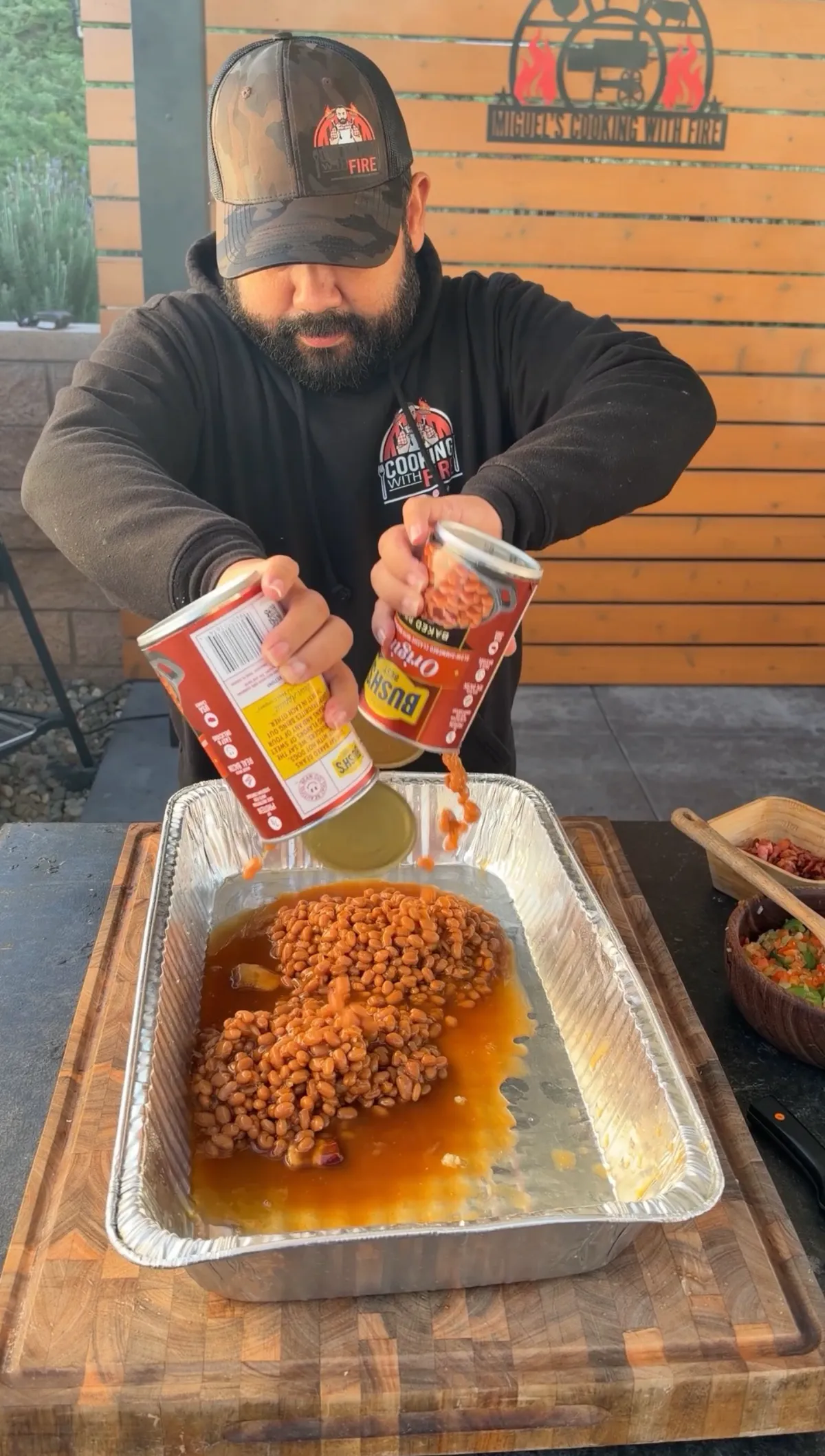 Man pouring two 55-ounce cans of Bush's Baked Beans into a large foil pan on a wooden cutting board