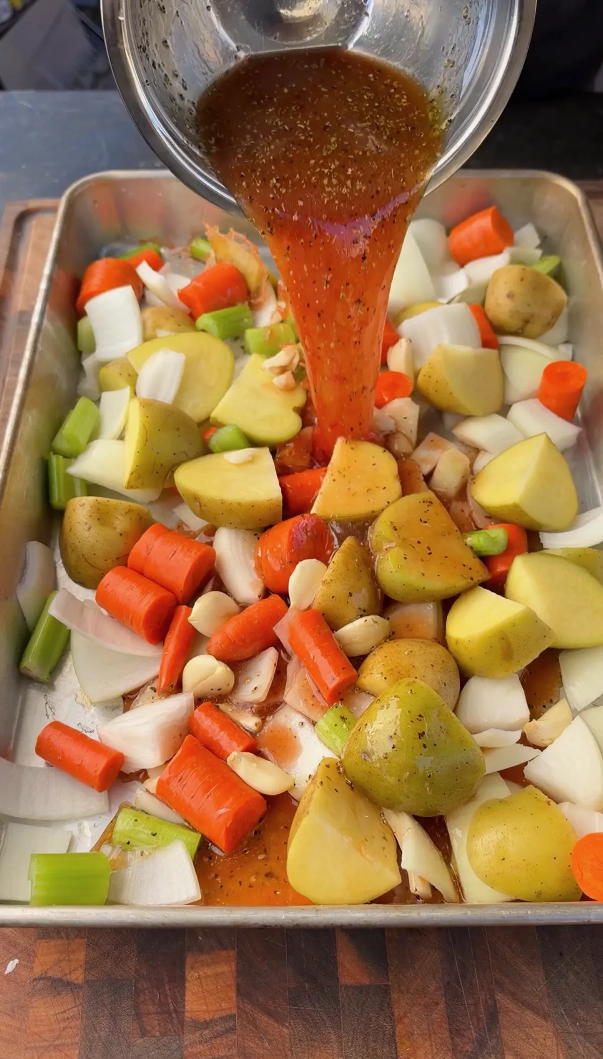 Braising liquid of beef broth, white wine, and lemon being poured over Yukon Gold potatoes, carrots, onions, celery, and garlic in a roasting tray