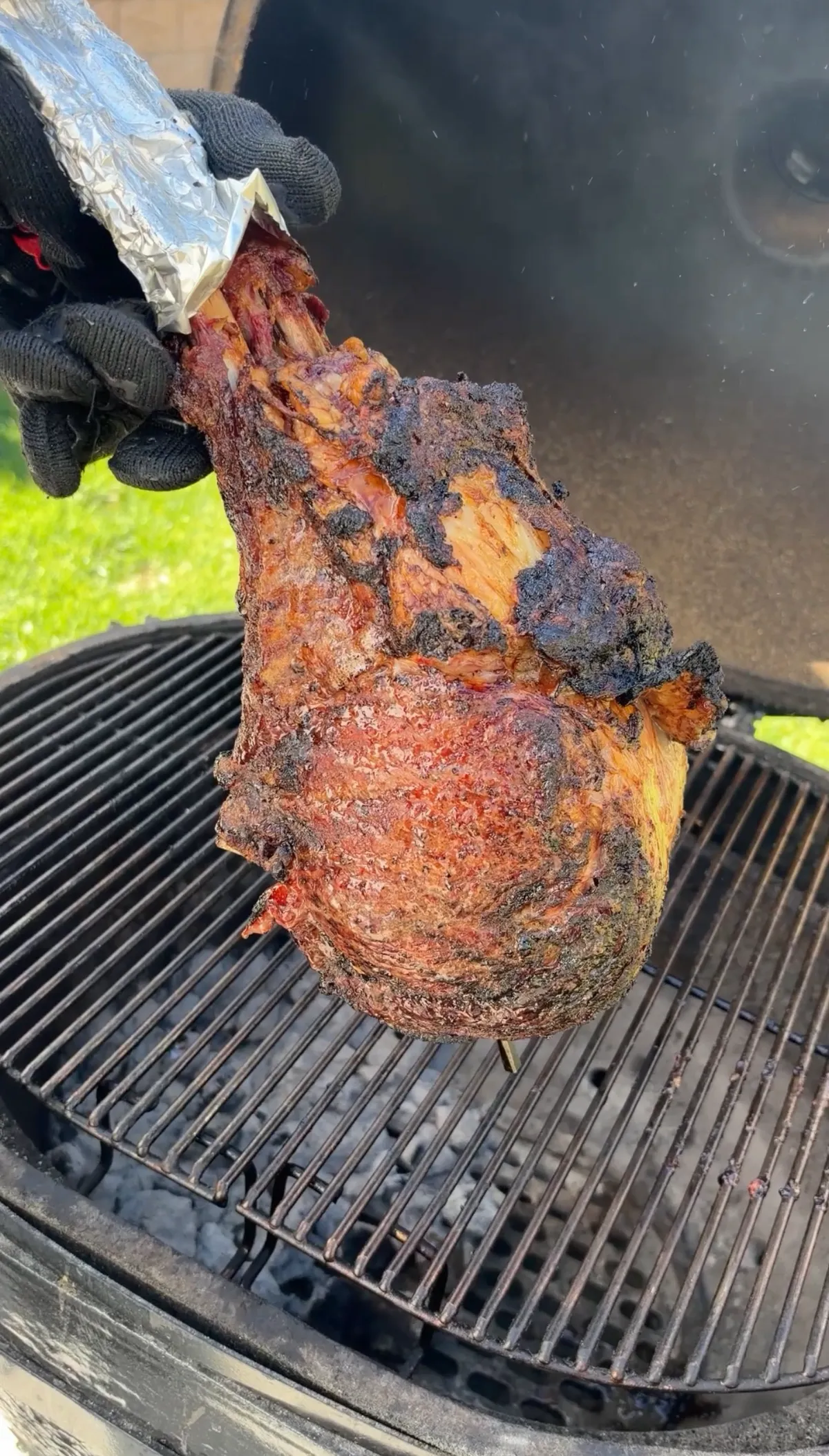 Two-bone prime rib tomahawk with deep bark crust being lifted by gloved hands from kamado grill grates