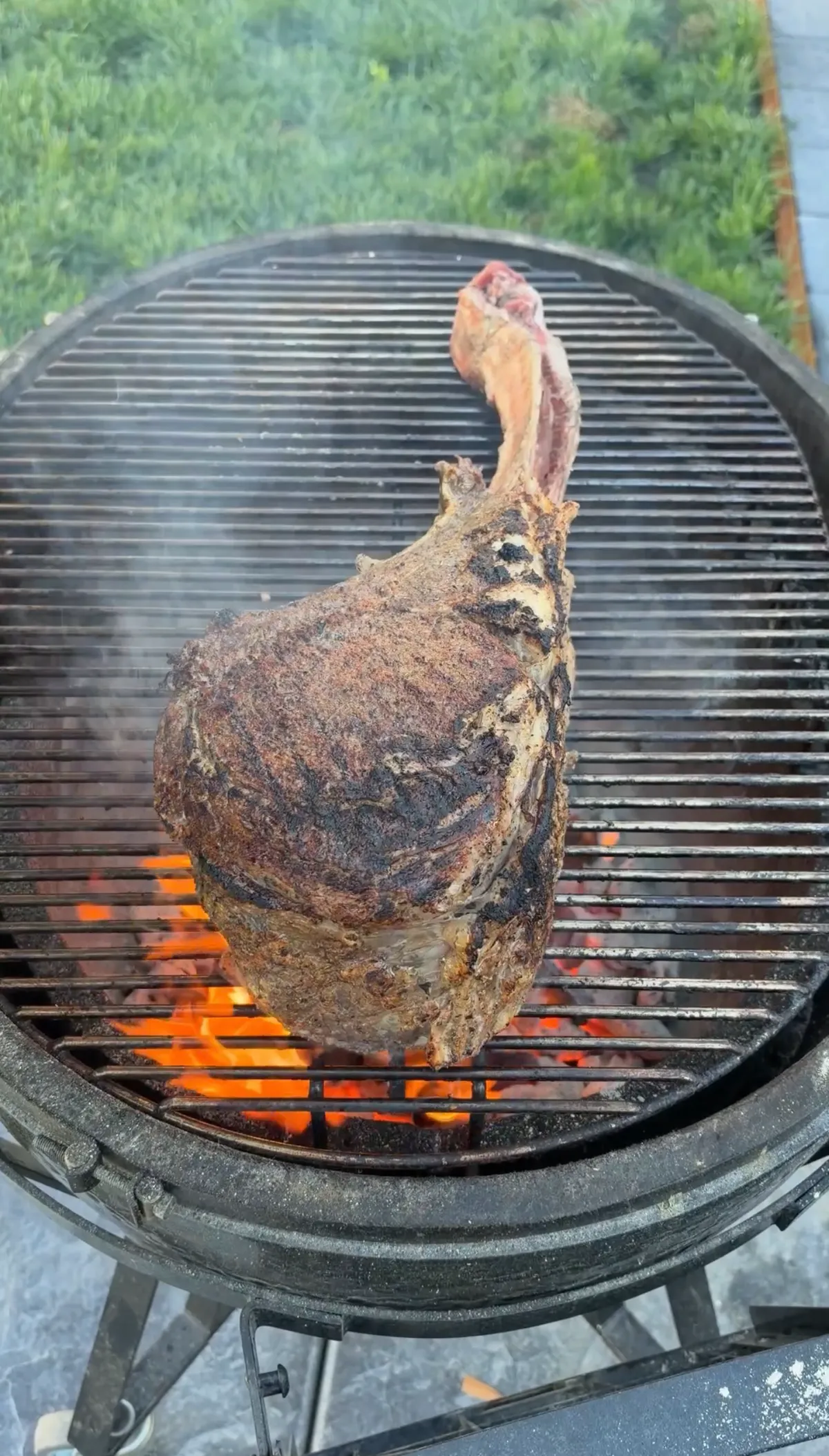Two-bone prime rib tomahawk standing upright on a kamado grill over open flames, developing a deep crust