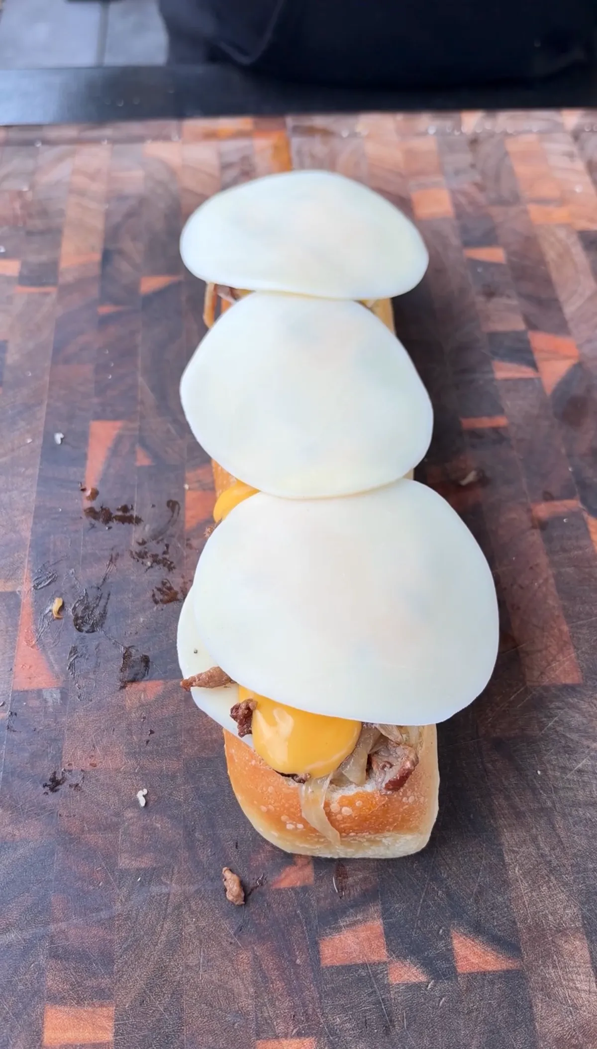 Top-down view of cheesesteak stuffed bread boat topped with overlapping melty provolone slices on a wooden cutting board