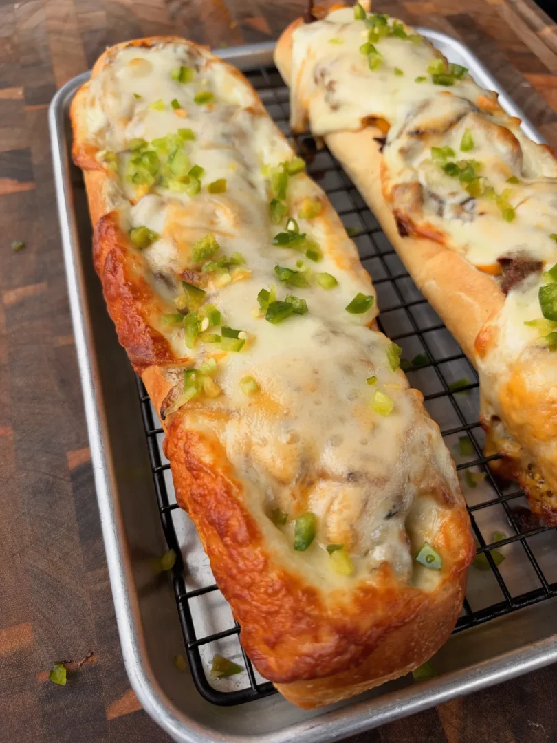 Cheesesteak stuffed bread boat topped with melted provolone and diced jalapeños on a wire rack baking sheet