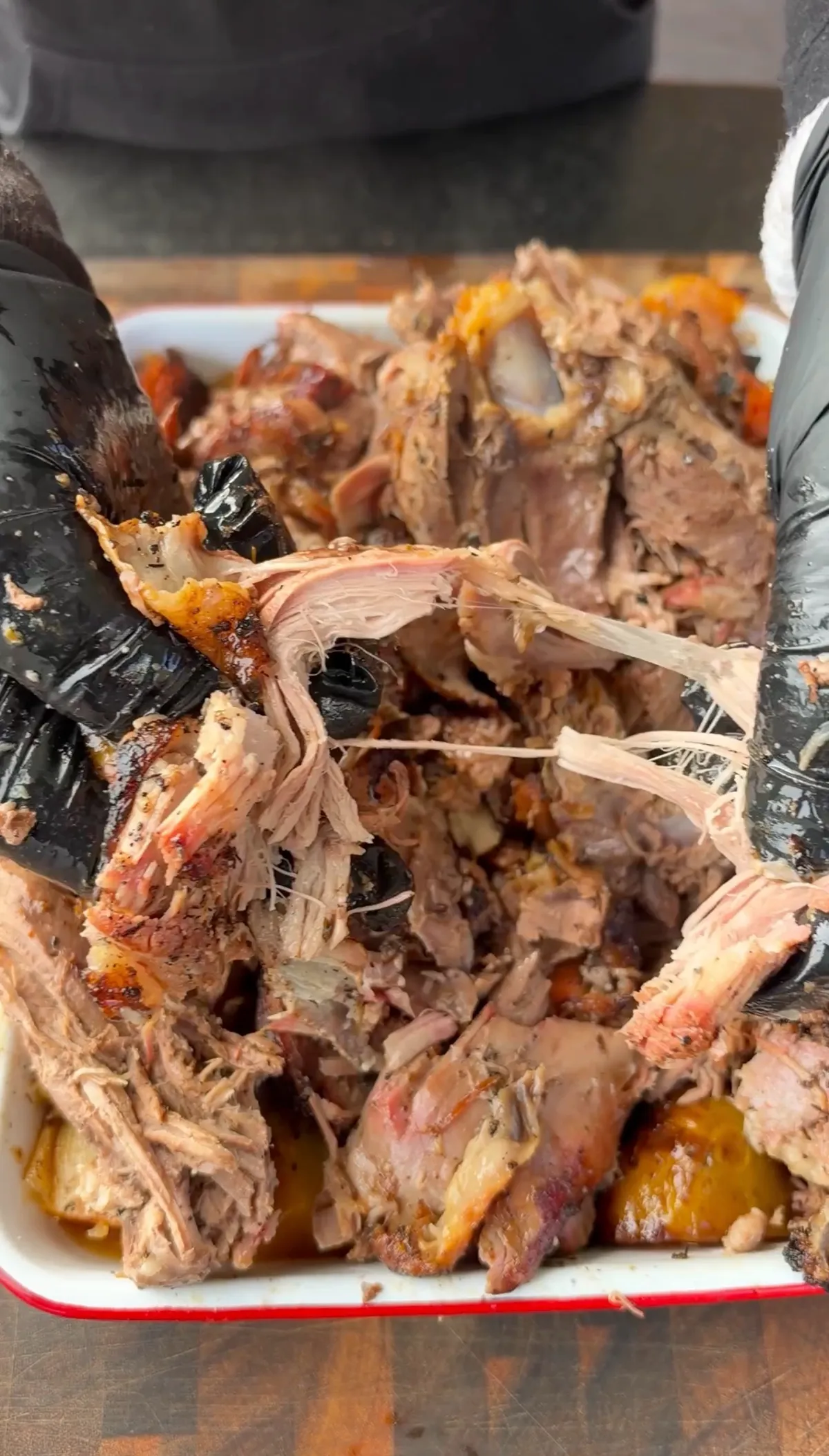 Hands in black gloves pulling apart fall-apart tender shredded Greek braised leg of lamb in white enamel roasting tray
