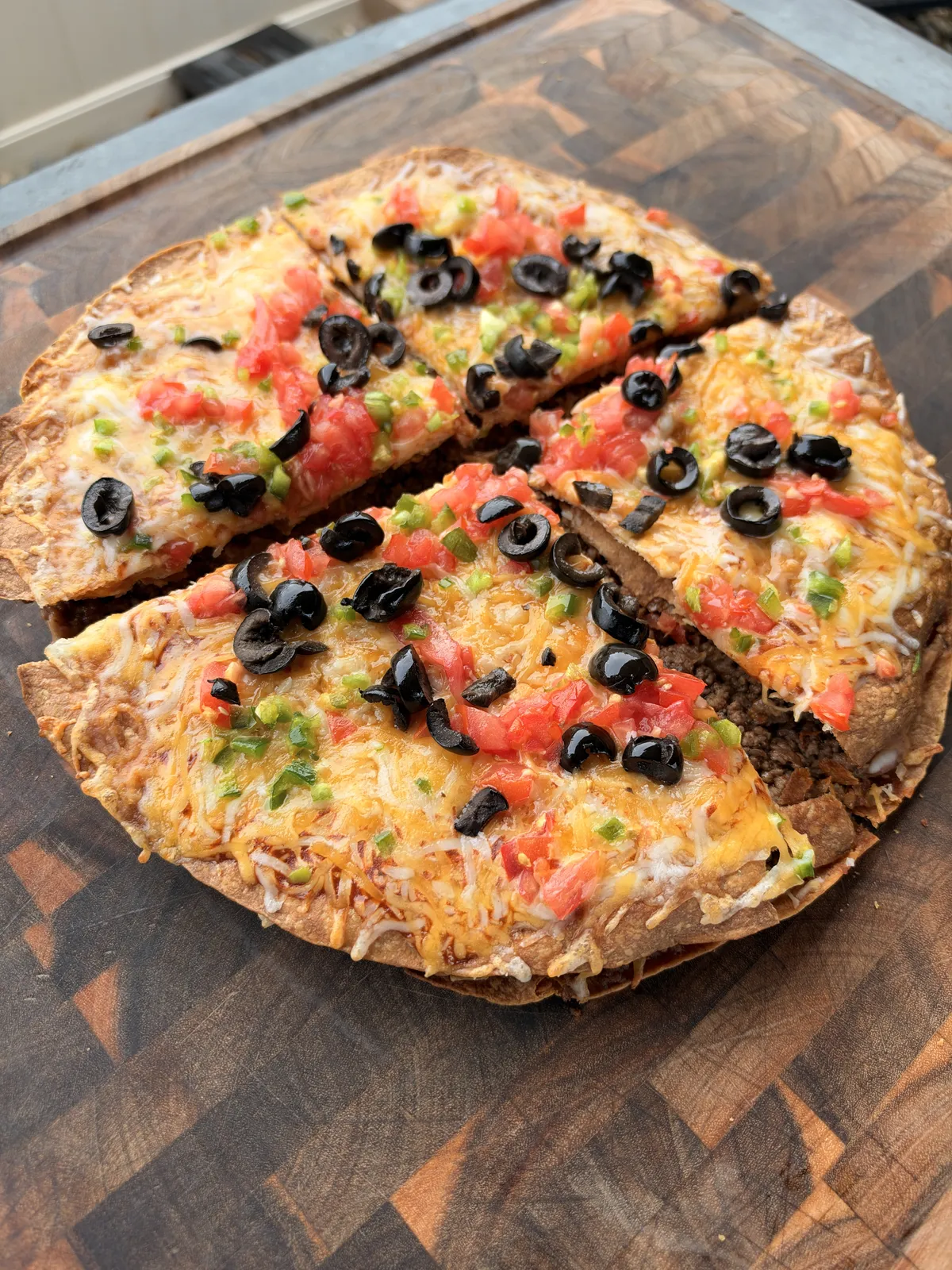 Taco Bell copycat Mexican pizza sliced into quarters on a wooden board, topped with melted Colby Jack cheese, diced tomatoes, sliced black olives, and jalapeños