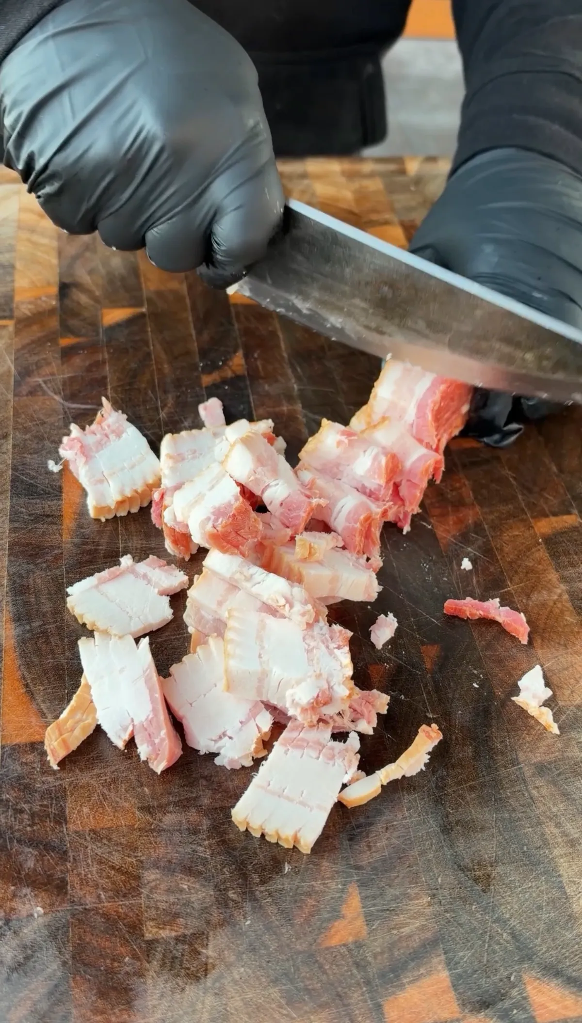 Hands in black gloves cutting bacon into ¼-inch pieces on a wooden cutting board with a chef's knife