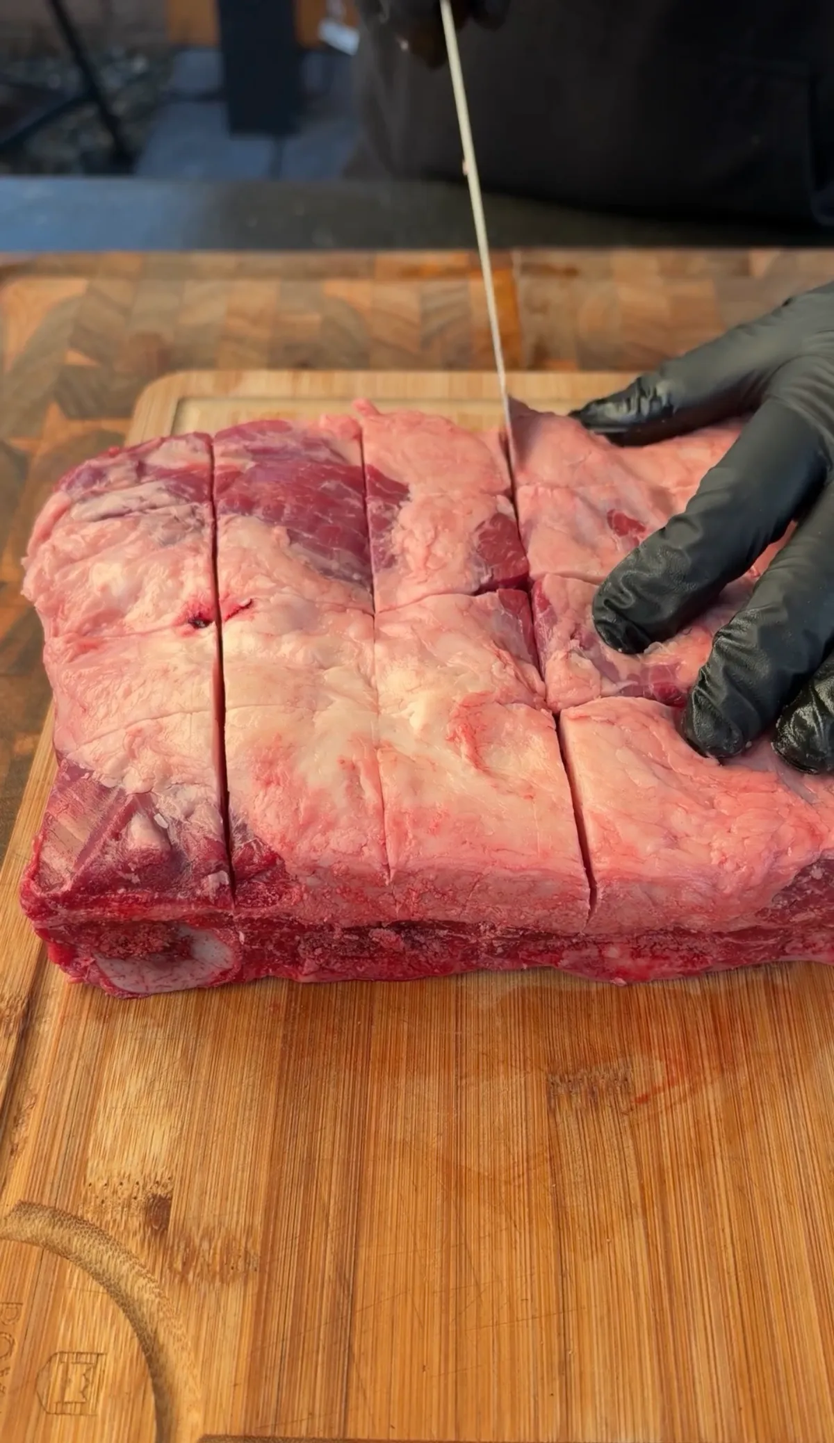 Gloved hands scoring a raw beef short rib slab with parallel cuts on a wooden cutting board