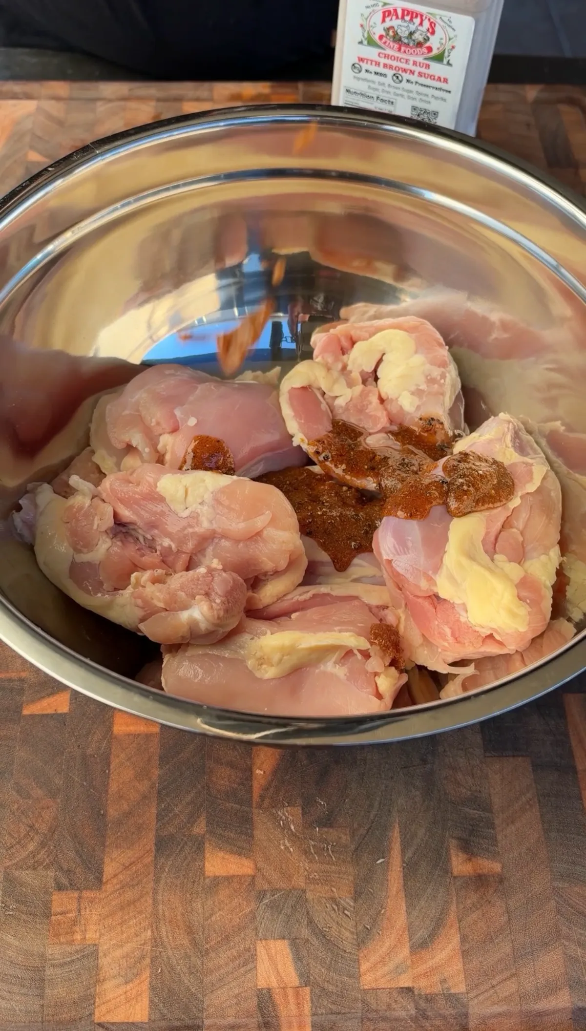 Raw boneless skinless chicken thighs in a stainless steel bowl with poultry seasoning on a wood cutting board