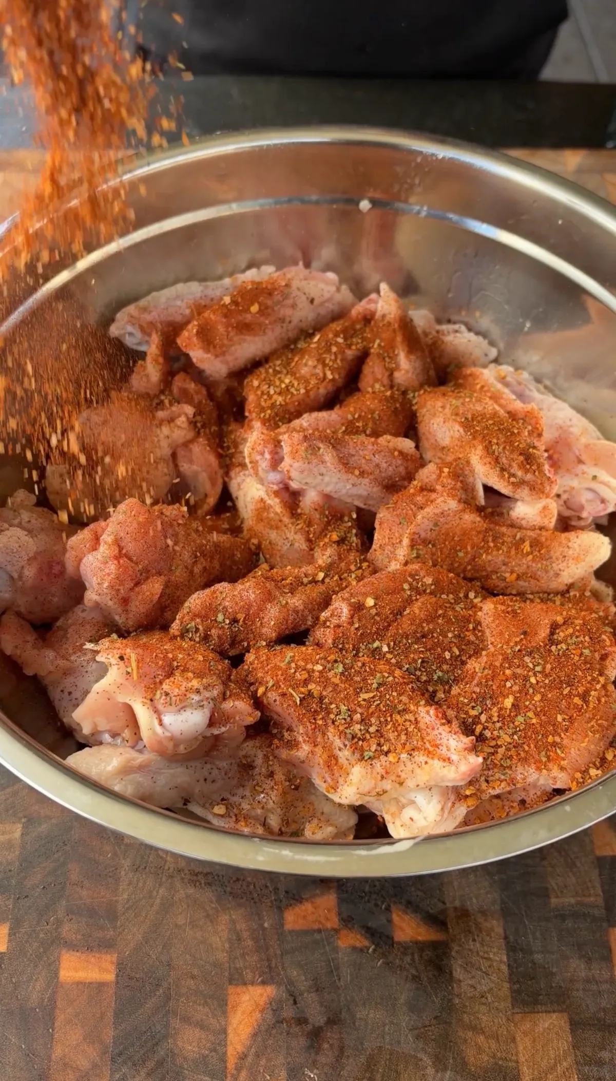 Raw chicken wings being generously coated with poultry seasoning in a large stainless steel mixing bowl