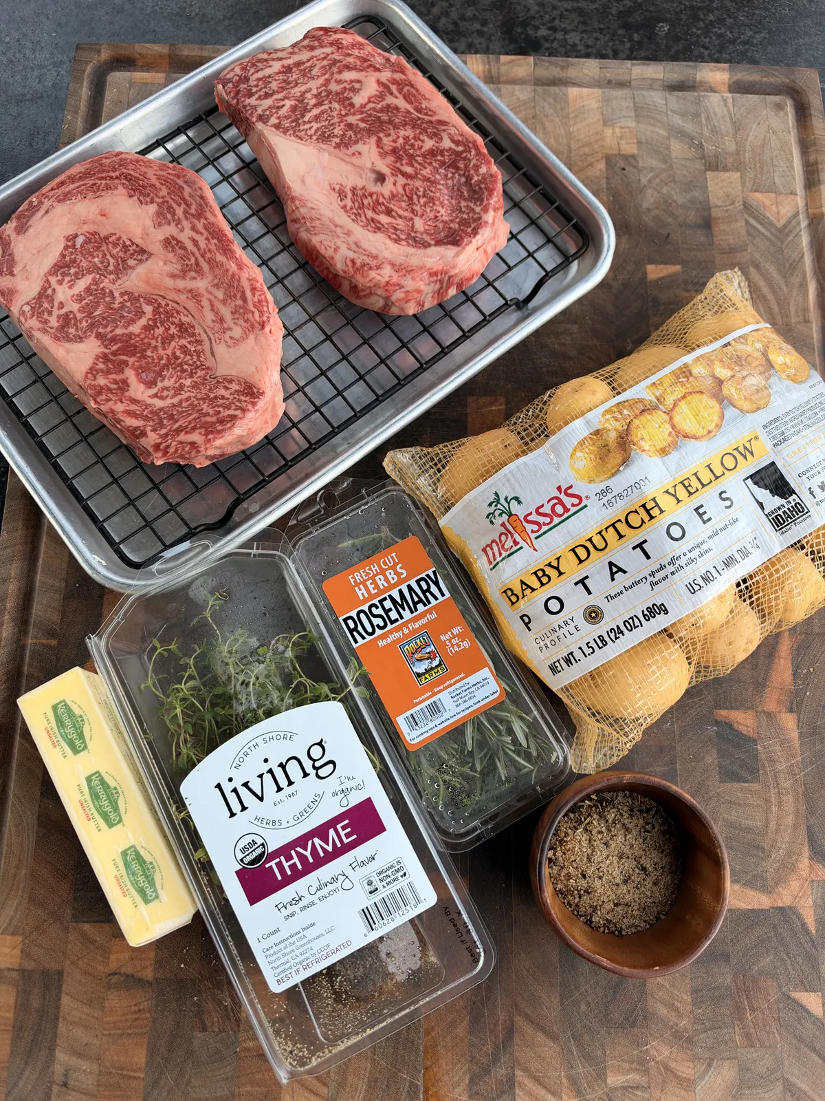 Raw marbled ribeye steaks on a wire rack with Kerrygold butter, fresh thyme, rosemary, steak seasoning, and baby Dutch yellow potatoes