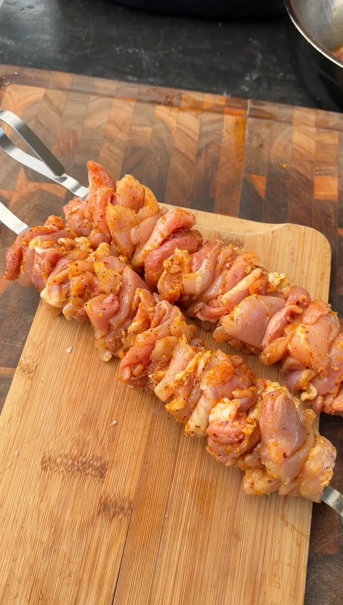 Raw boneless chicken thighs seasoned with poultry seasoning and threaded onto metal skewers on a wooden cutting board