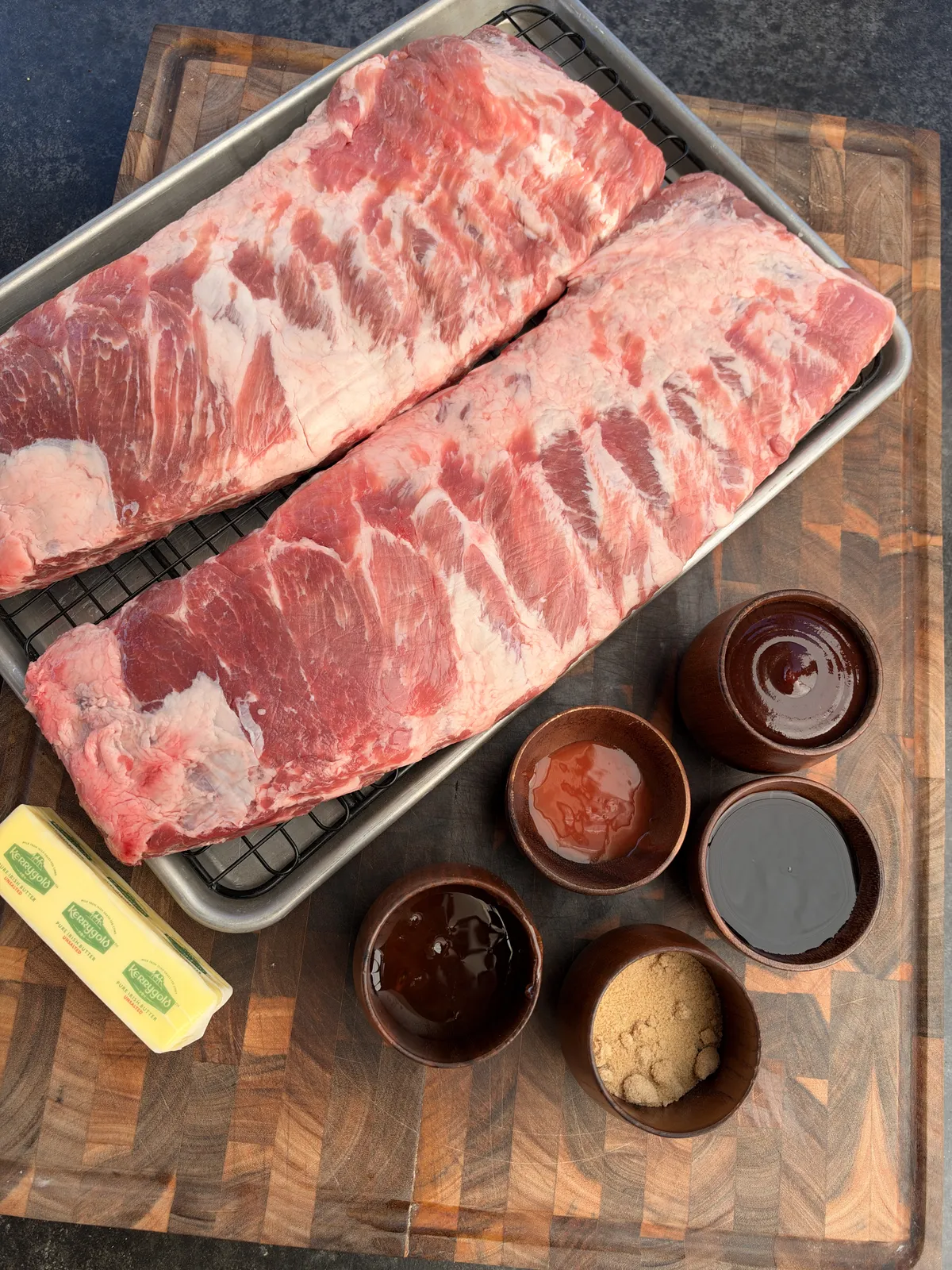 Two raw St. Louis-style pork rib racks on a tray with butter, honey, soy sauce, brown sugar, and barbecue sauce