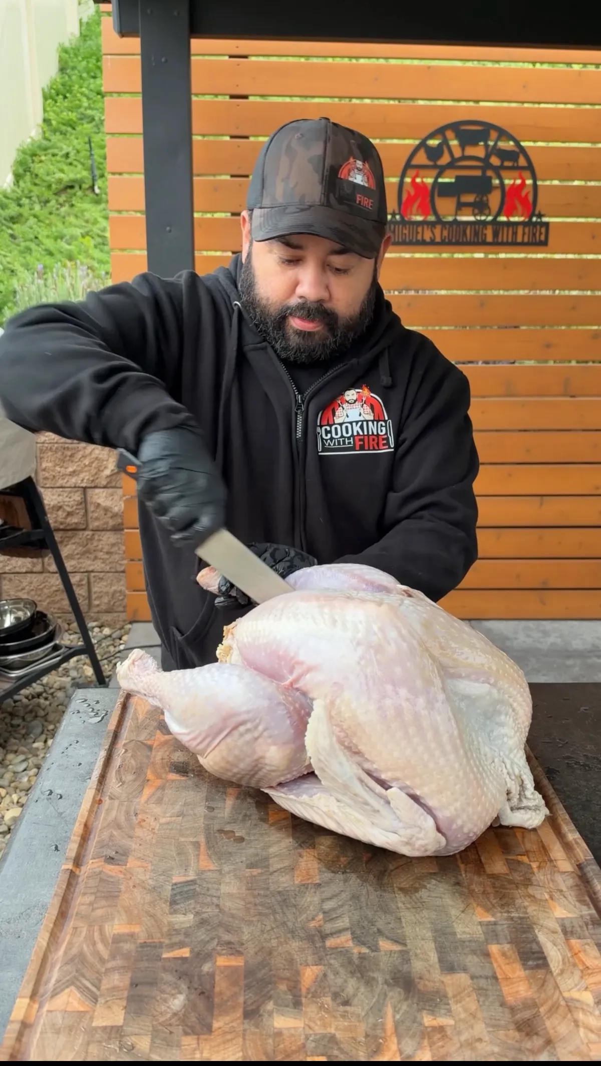 Bearded man in Cooking with Fire hoodie cutting whole raw turkey frog-style on wood cutting board