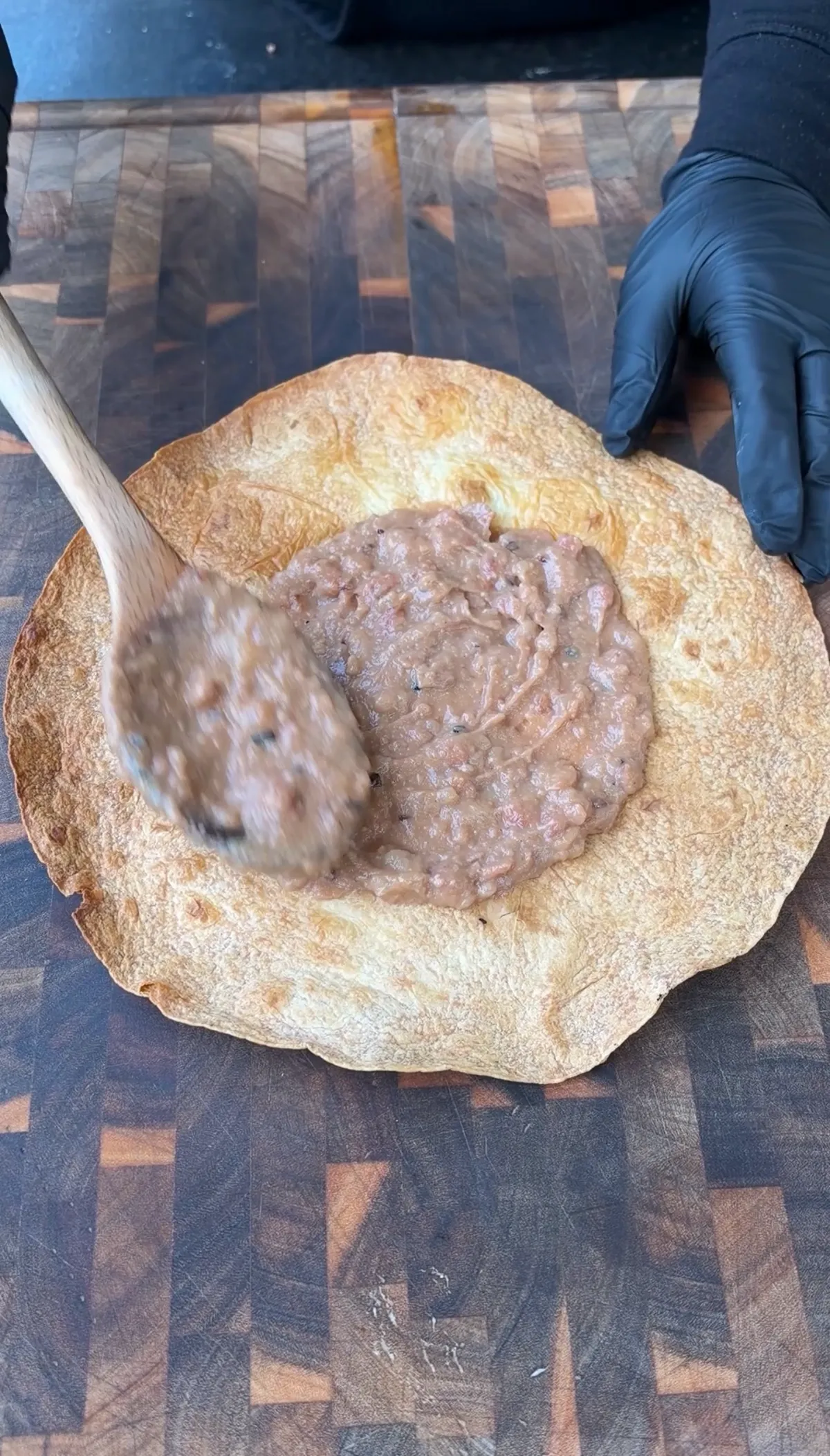Spreading seasoned refried beans onto a crispy flour tortilla with a wooden spoon on a wood cutting board