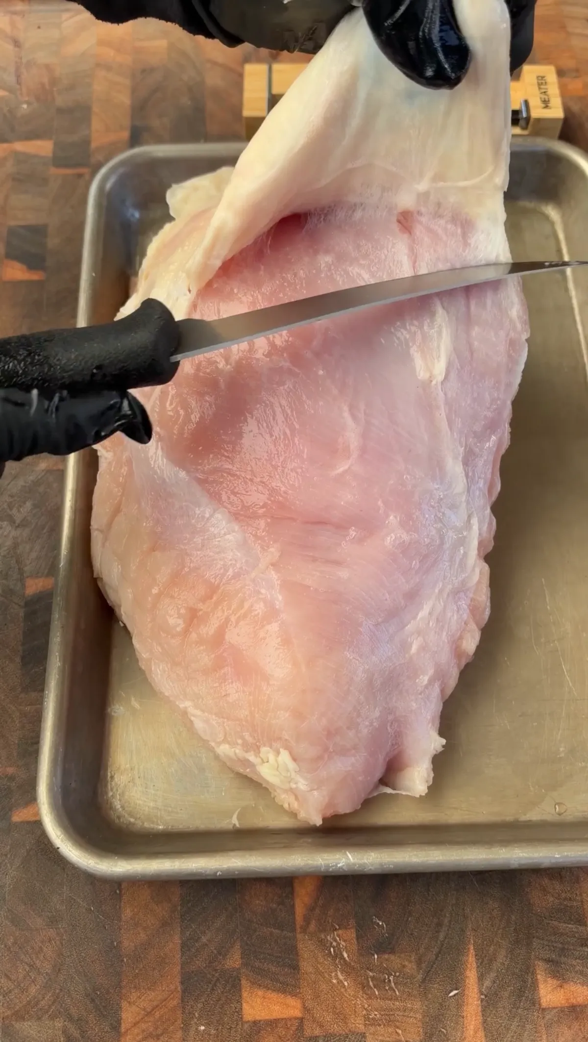 Gloved hands using a sharp knife to remove skin from a raw boneless turkey breast on a metal tray