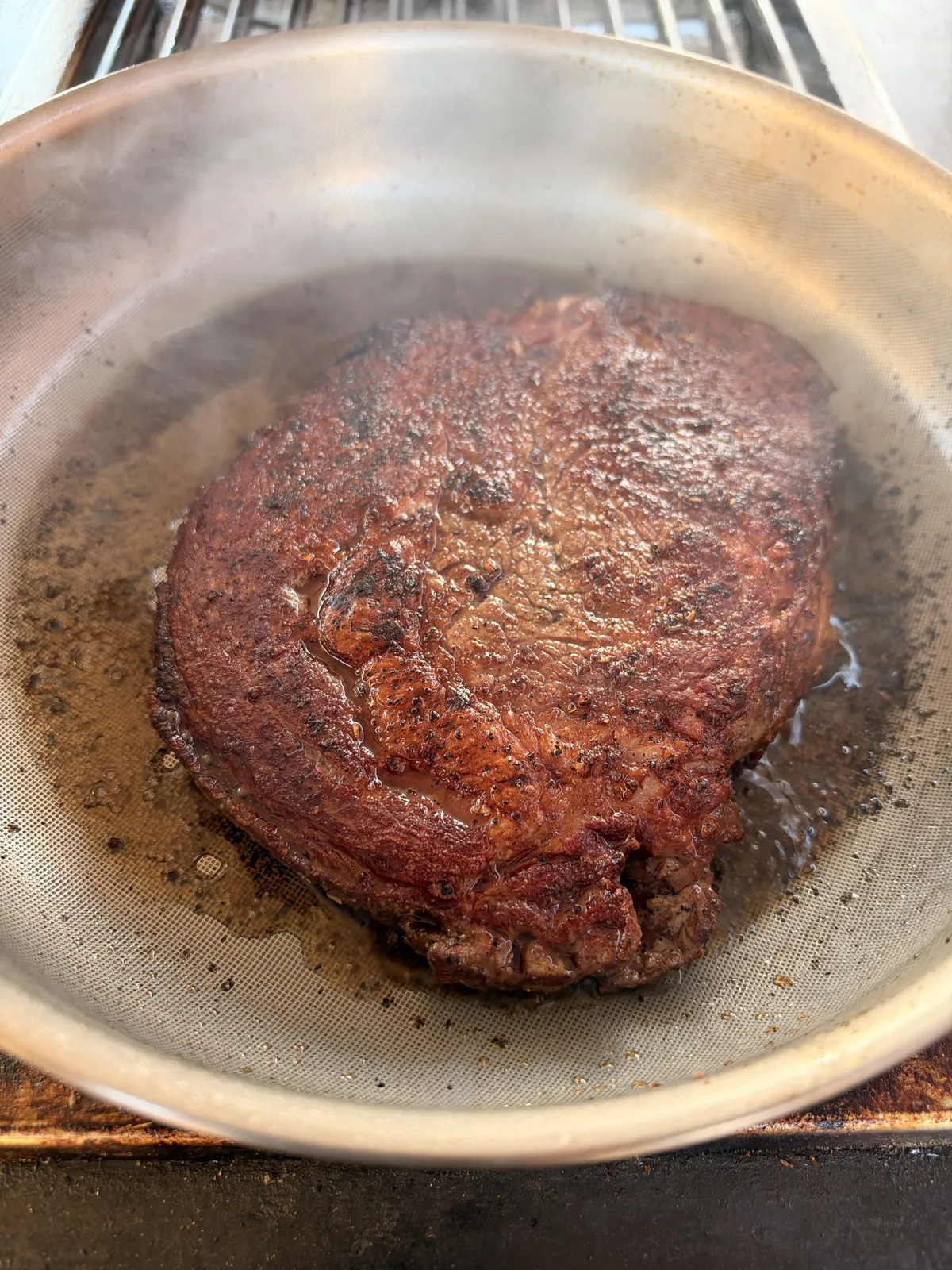 Ribeye steak sizzling in a stainless steel pan with steam rising during the garlic herb butter basting step