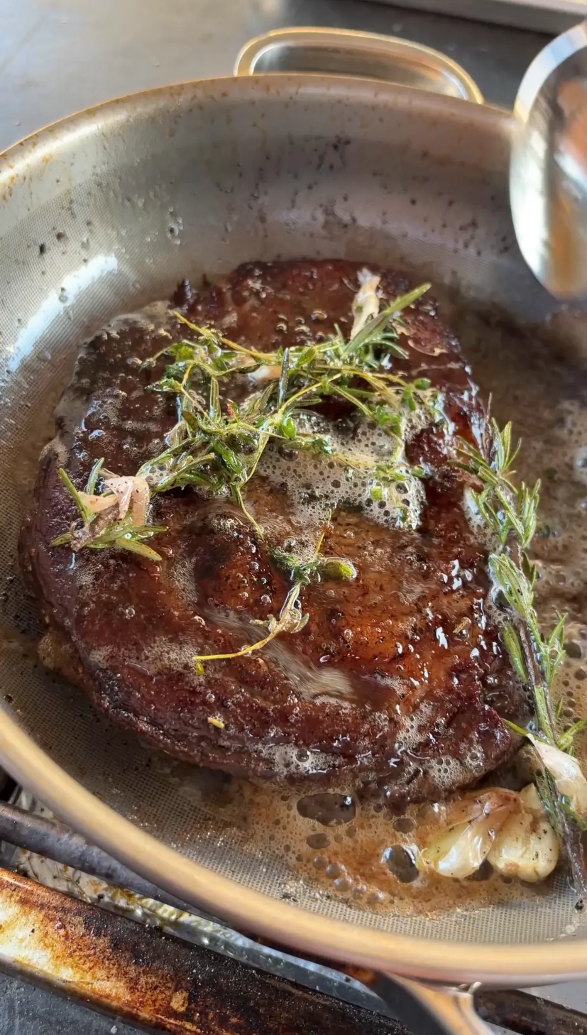 Ribeye steak basting in foamy garlic herb butter with rosemary and thyme sprigs in a stainless pan