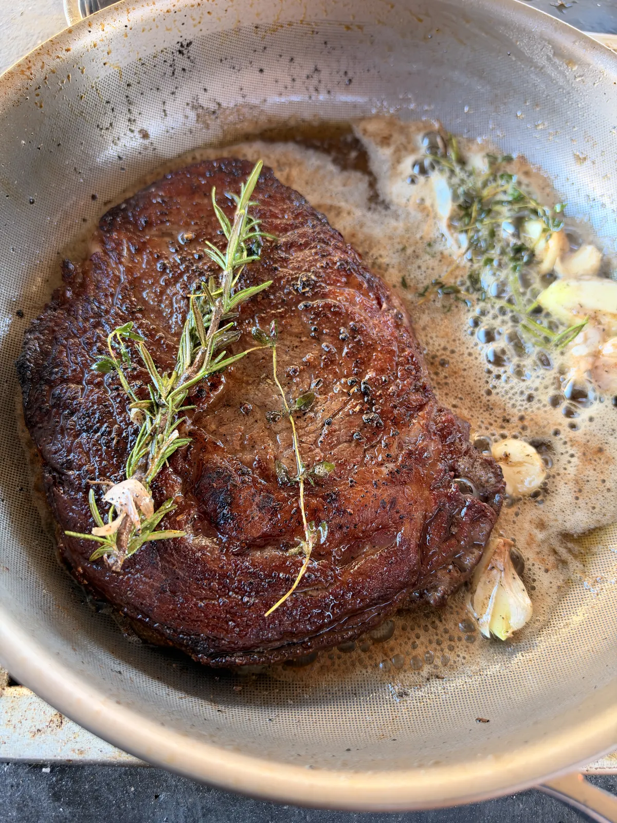 Pan-seared ribeye steak topped with rosemary and thyme sprigs in garlic herb butter with smashed garlic cloves