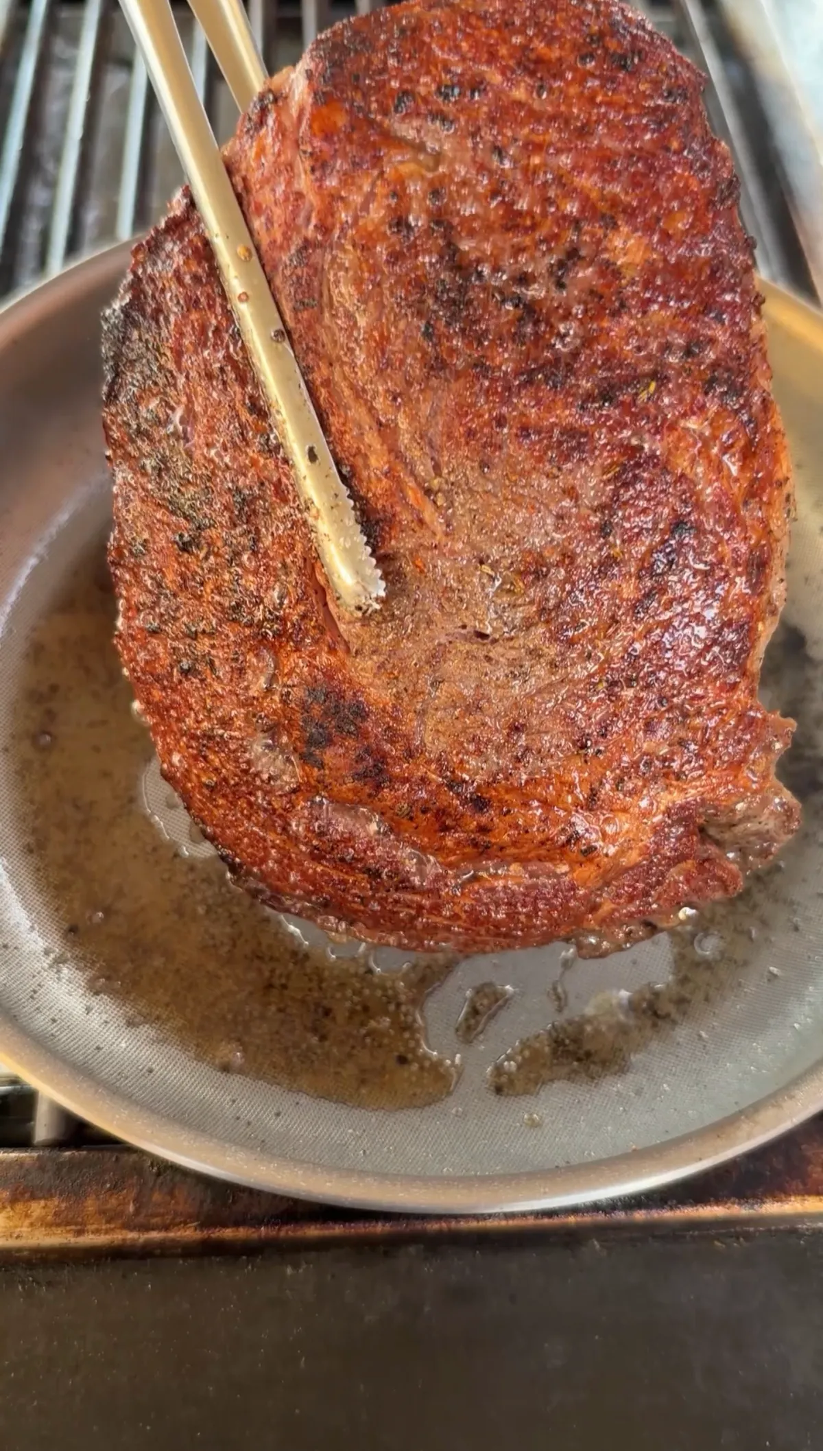 Ribeye steak held upright on its edge with tongs in a stainless pan, showing deep golden-brown seared crust
