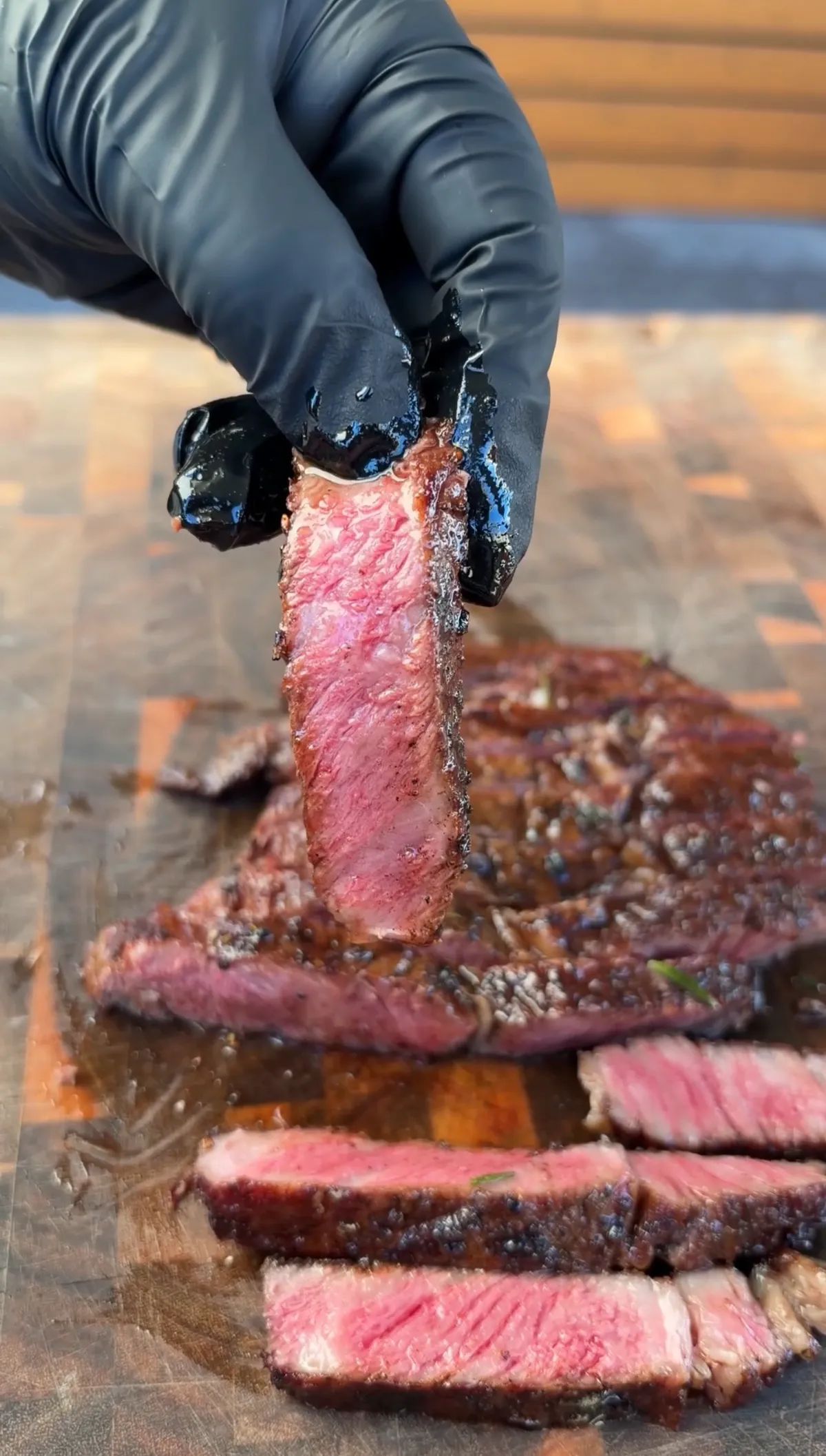 Gloved hand holding up a sliced ribeye piece showing pink medium-rare interior above sliced steak on cutting board