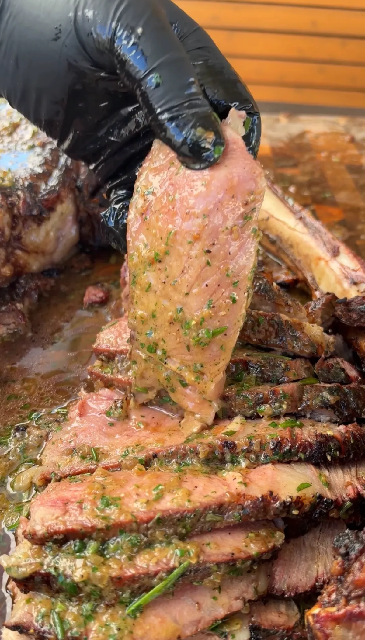 Gloved hand lifting sliced prime rib tomahawk with roasted garlic, parsley and herb compound butter pooling over the cuts
