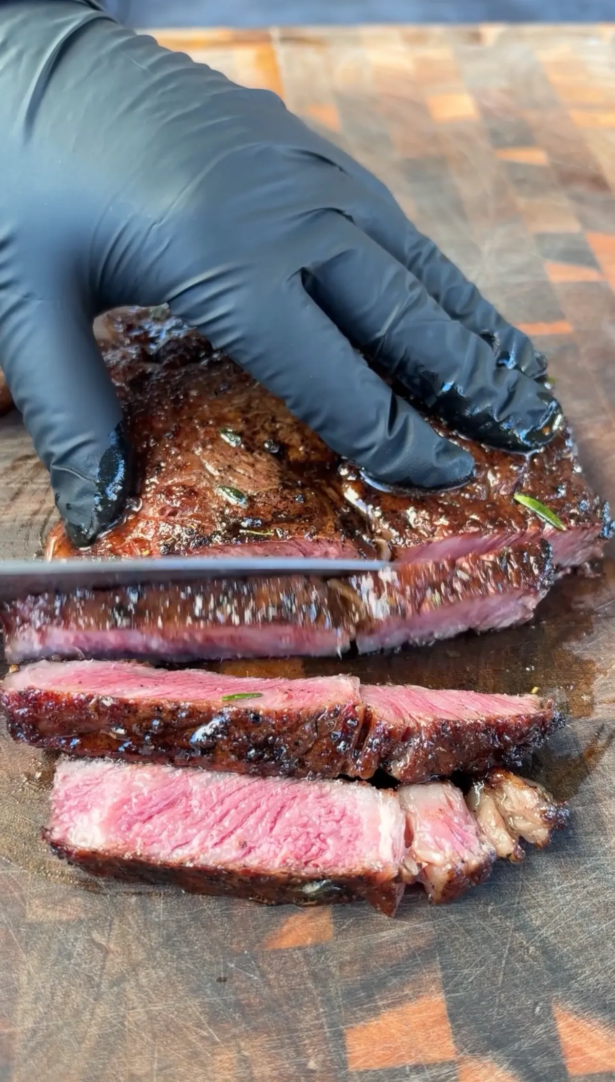 Gloved hands slicing a pan-seared ribeye against the grain on a wooden cutting board, revealing a pink medium-rare interior