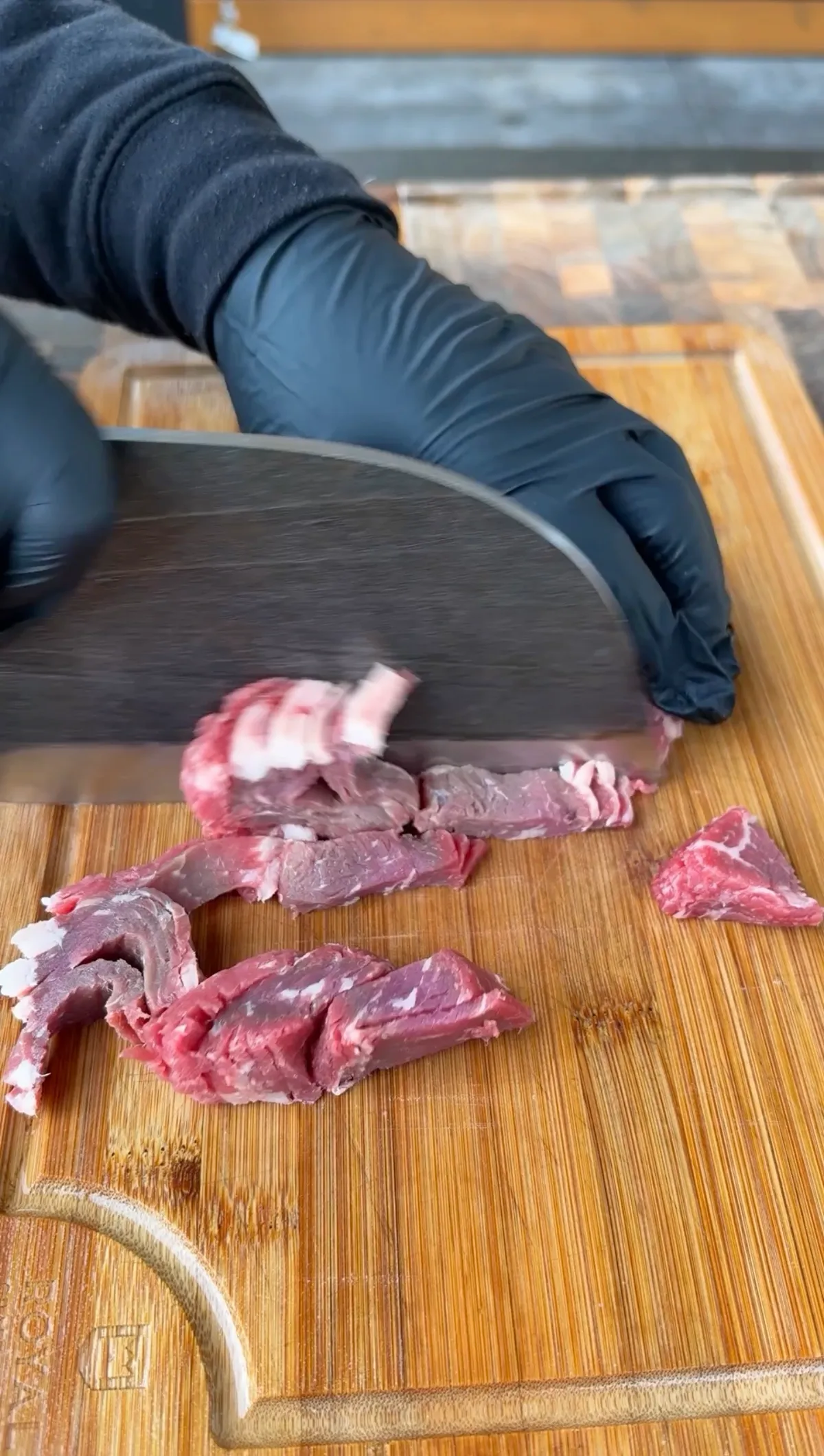 Gloved hands using a cleaver to thinly slice ribeye steak into strips on a wooden cutting board
