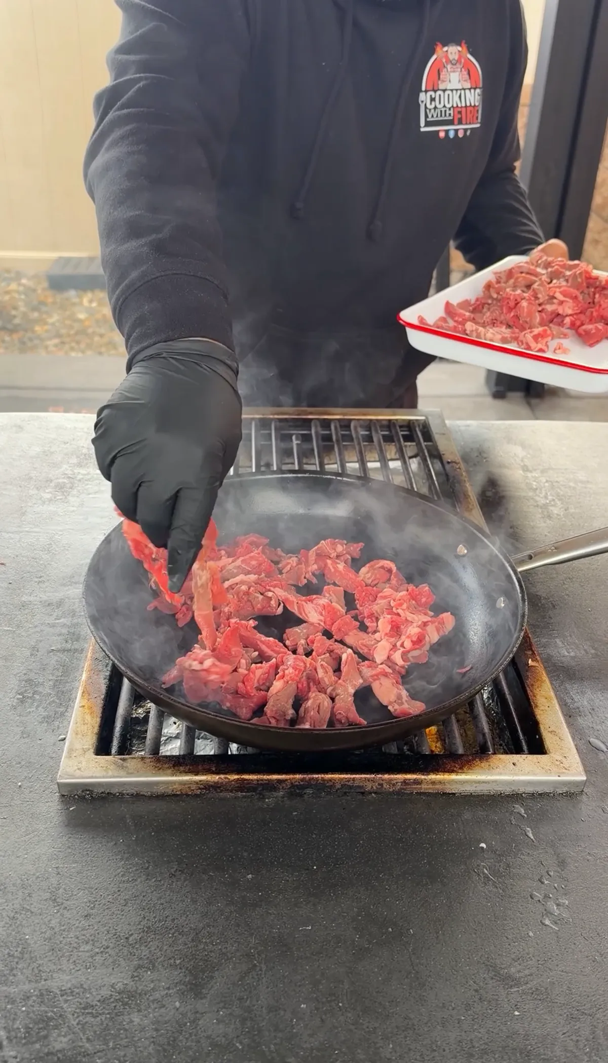 Thinly sliced ribeye steak strips searing in a smoking hot cast iron skillet on a grill grate