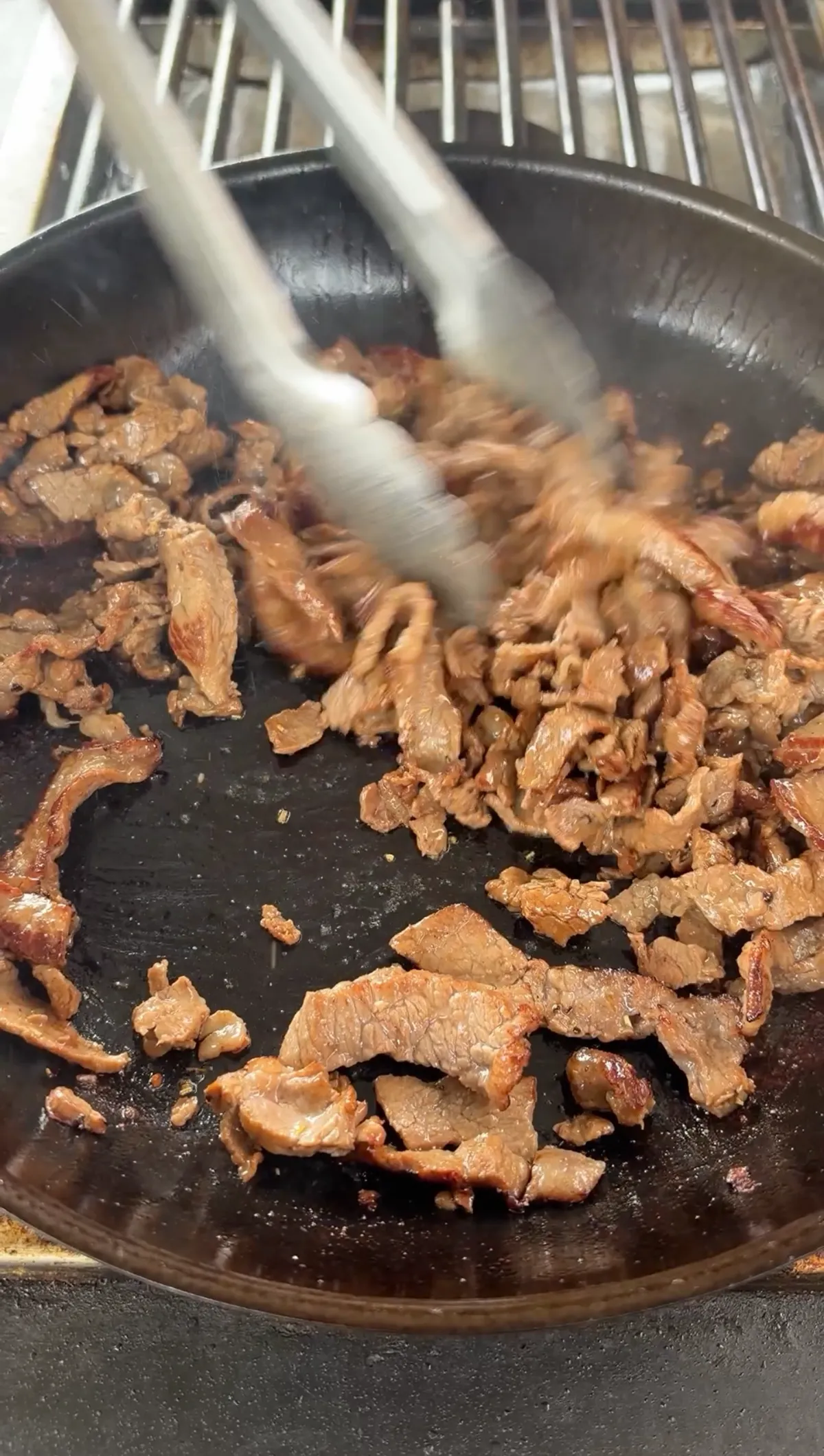 Thinly sliced ribeye steak strips being tossed with tongs in a hot skillet until browned and cooked through