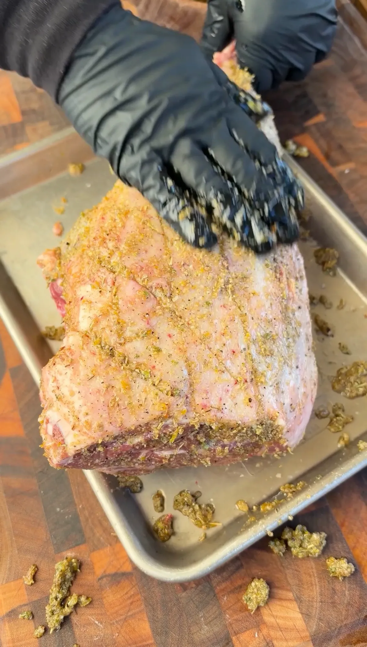 Gloved hands pressing Greek oregano and lemon zest seasoning mixture into scored leg of lamb on a baking sheet