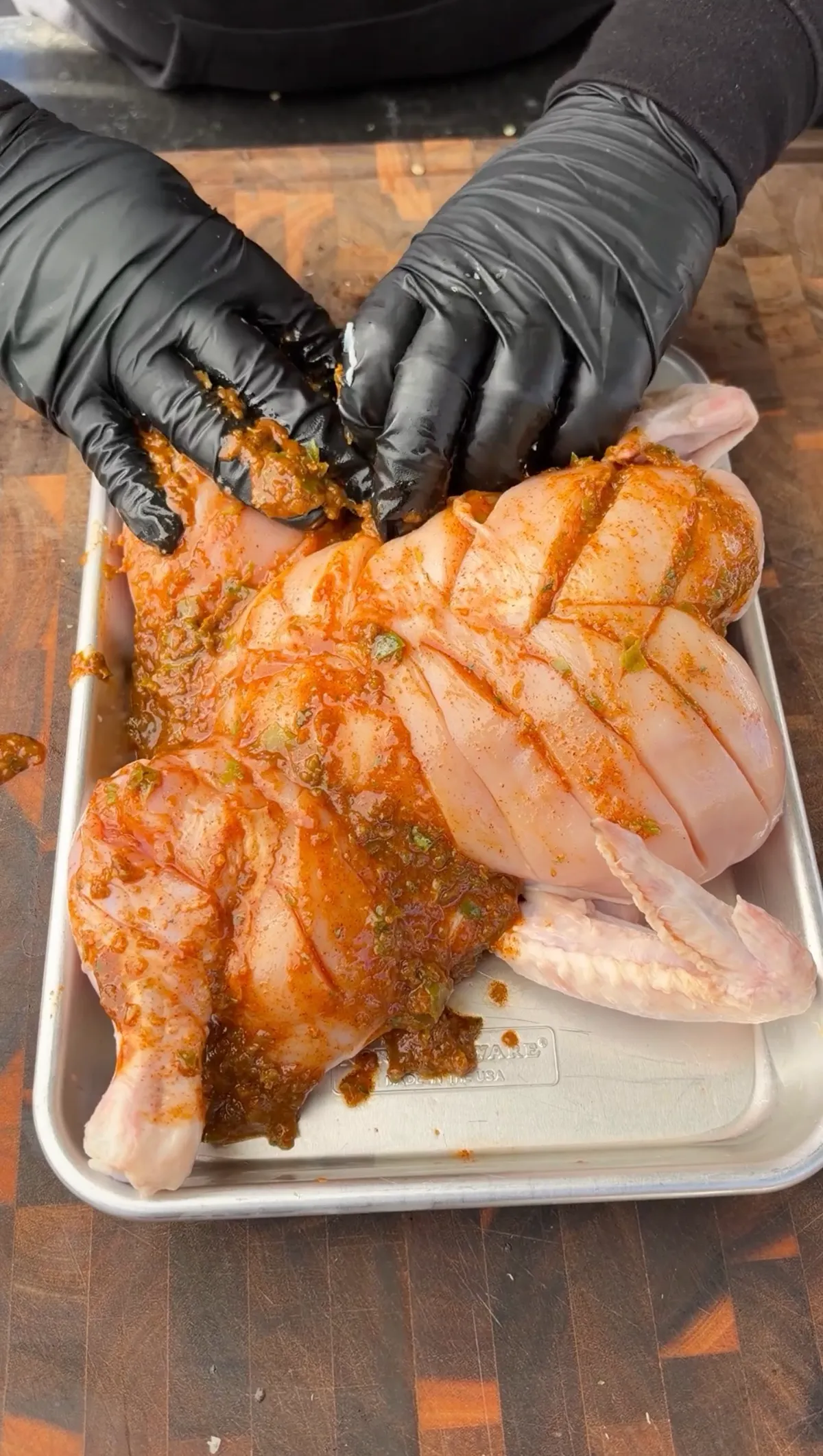 Gloved hands rubbing green chile lime marinade into scored spatchcocked chicken on a sheet pan