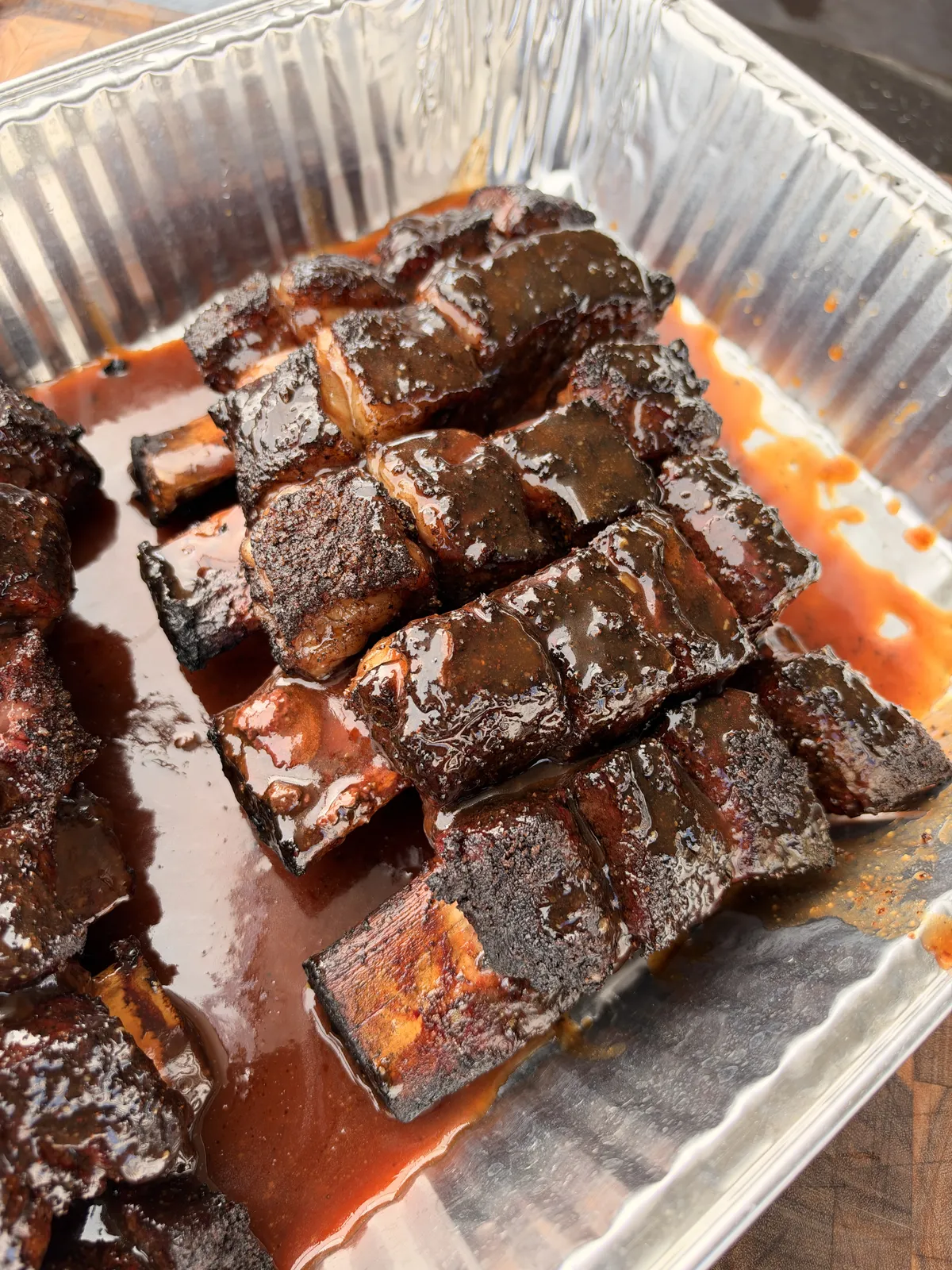 Glossy BBQ-glazed beef short ribs stacked in a disposable aluminum pan with rich red barbecue sauce pooling beneath