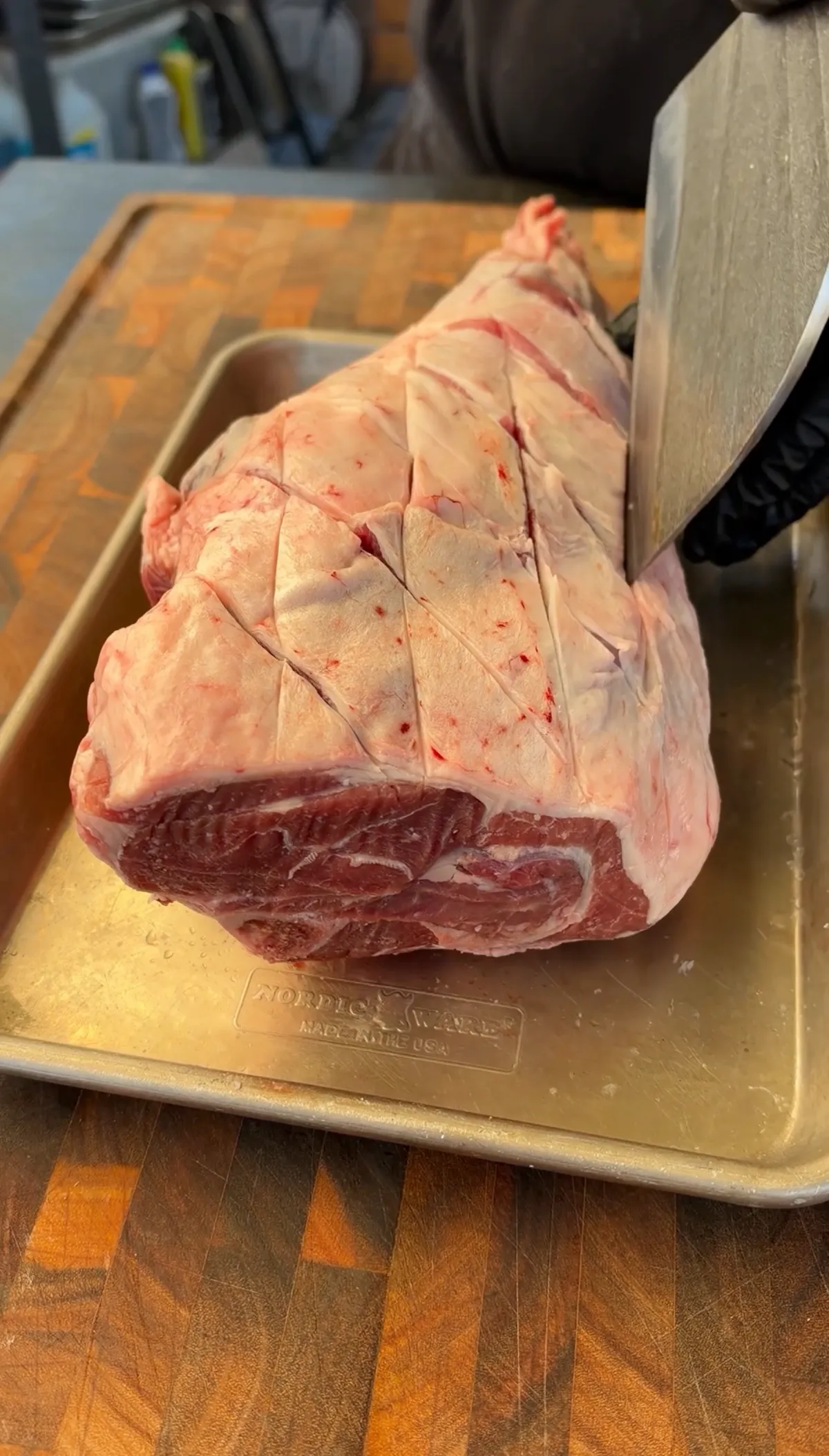 Sharp knife scoring a crosshatch pattern into the fat cap of a raw leg of lamb on a Nordic Ware baking sheet