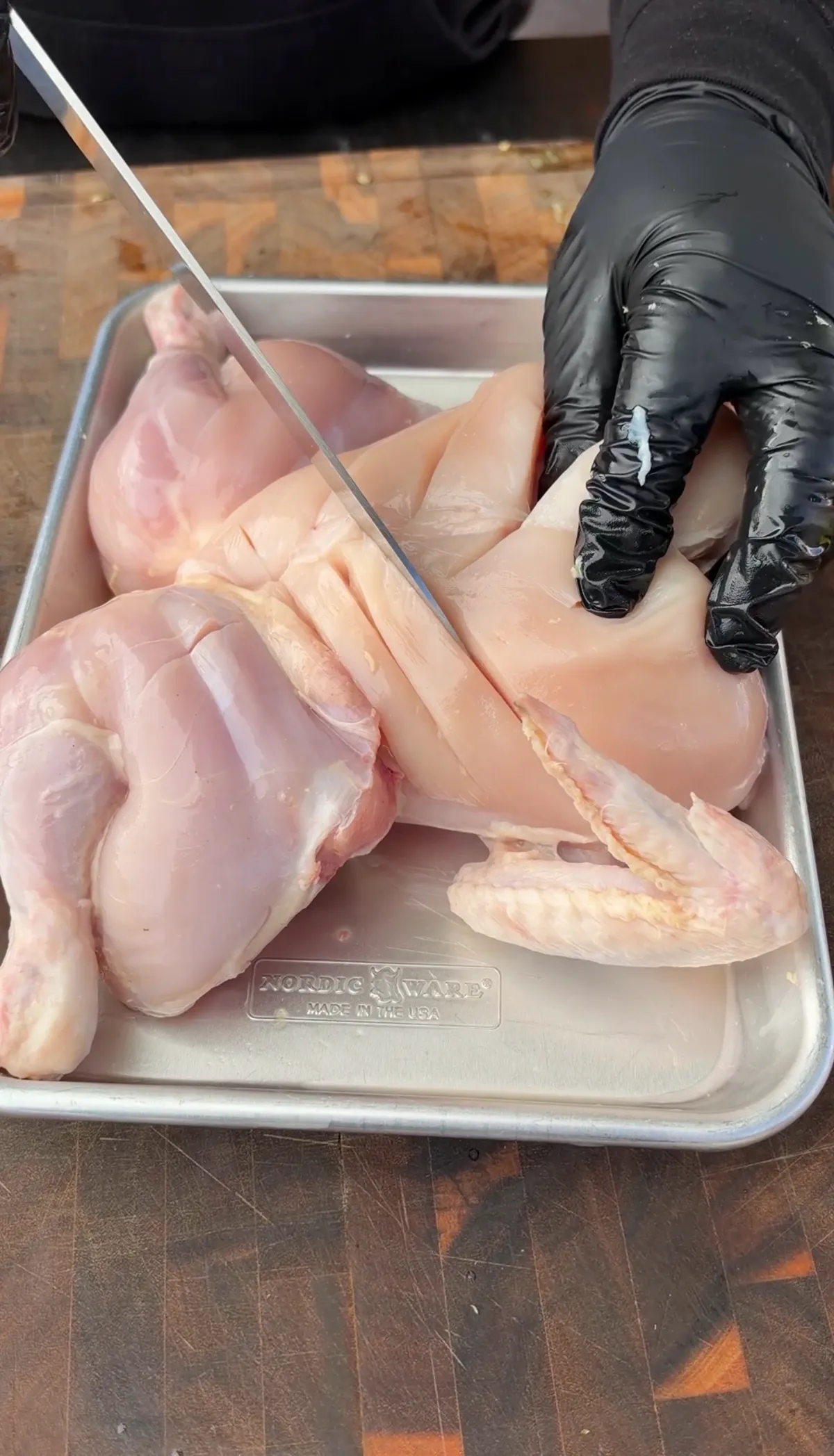 Gloved hands using a sharp knife to score a raw spatchcocked chicken in a Nordic Ware sheet pan